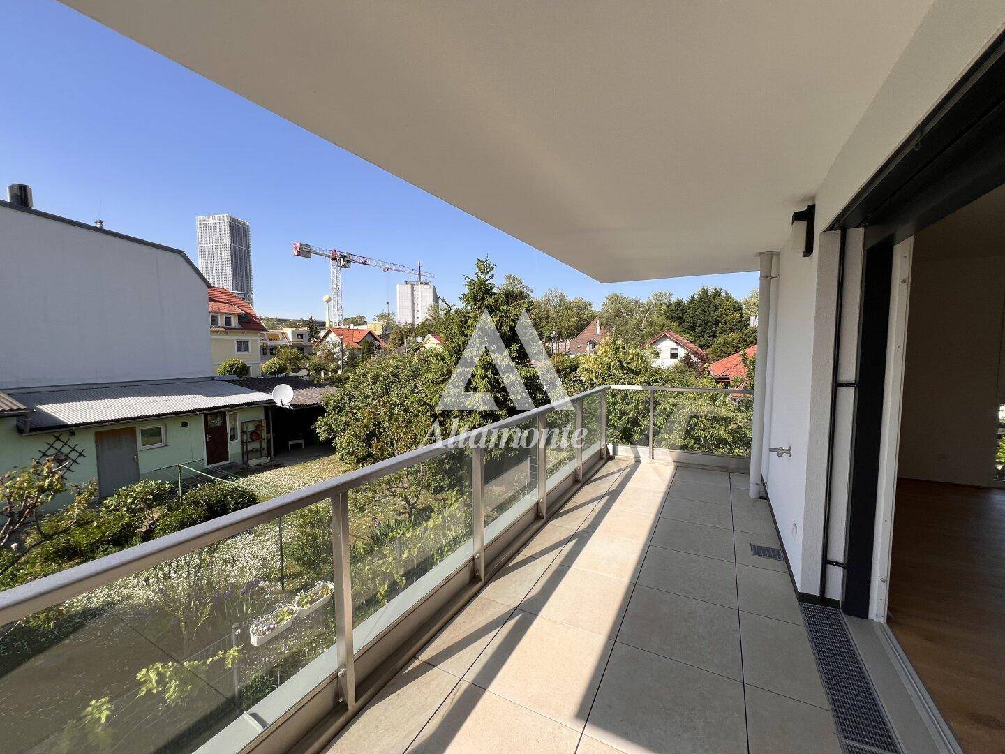 Langer Balkon mit Glasgeländer und Blick auf die umliegende Wohngegend und Bäume.