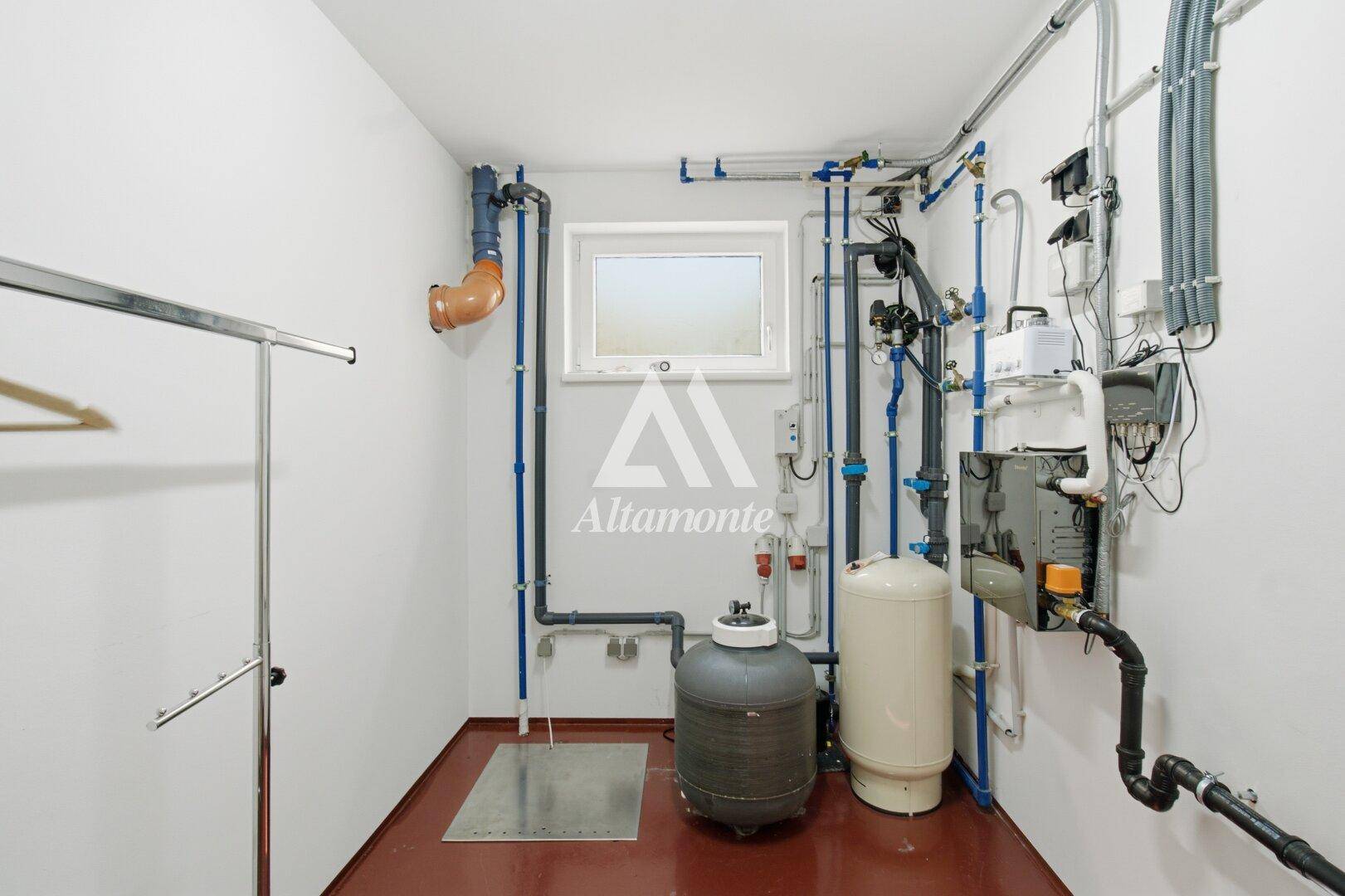 Technikraum mit zahlreichen Installationen, Rohren und einem kleinen Fenster.