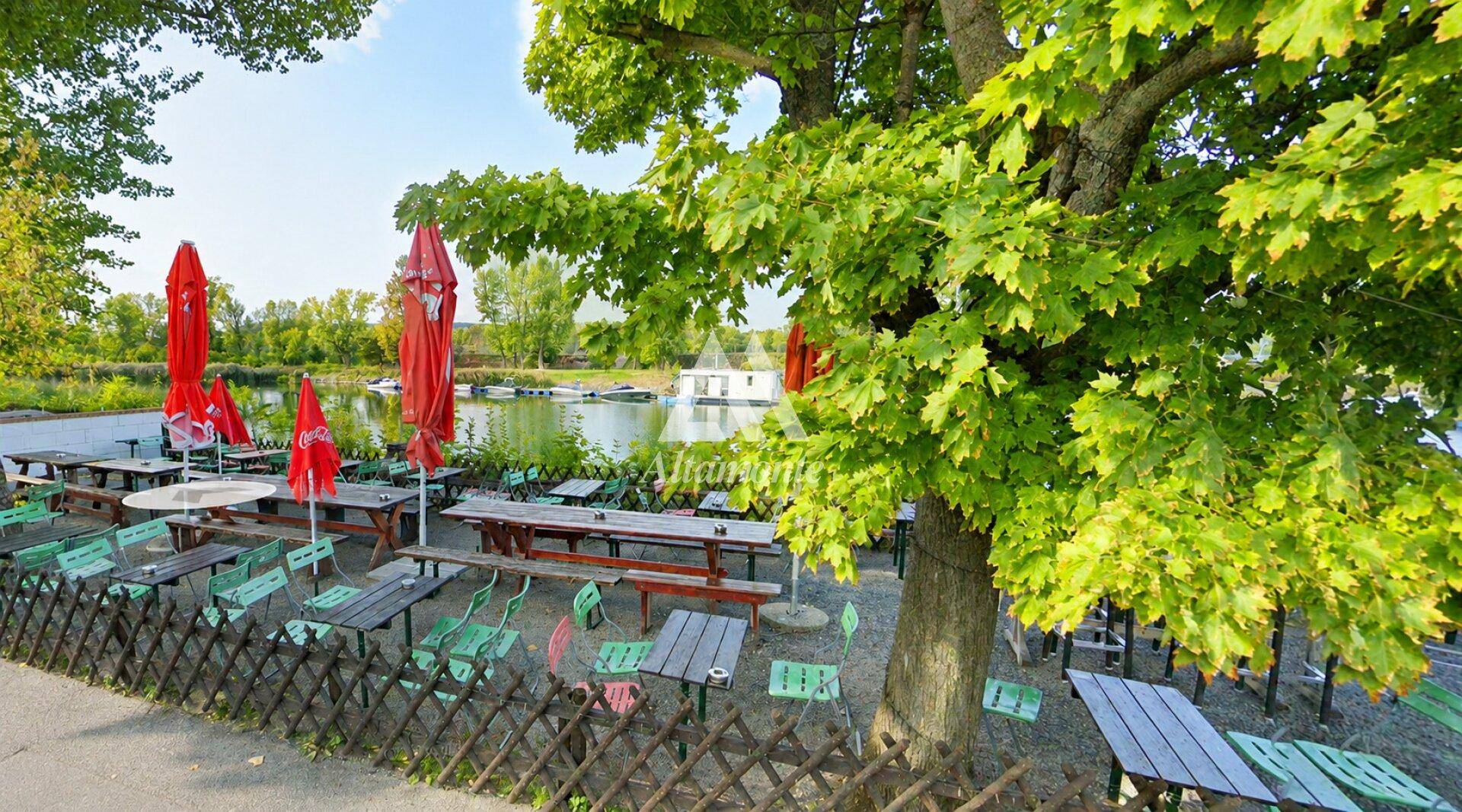 Großer Außenbereich mit zahlreichen Tischen und roten Sonnenschirmen direkt am Seeufer unter einem Baum.