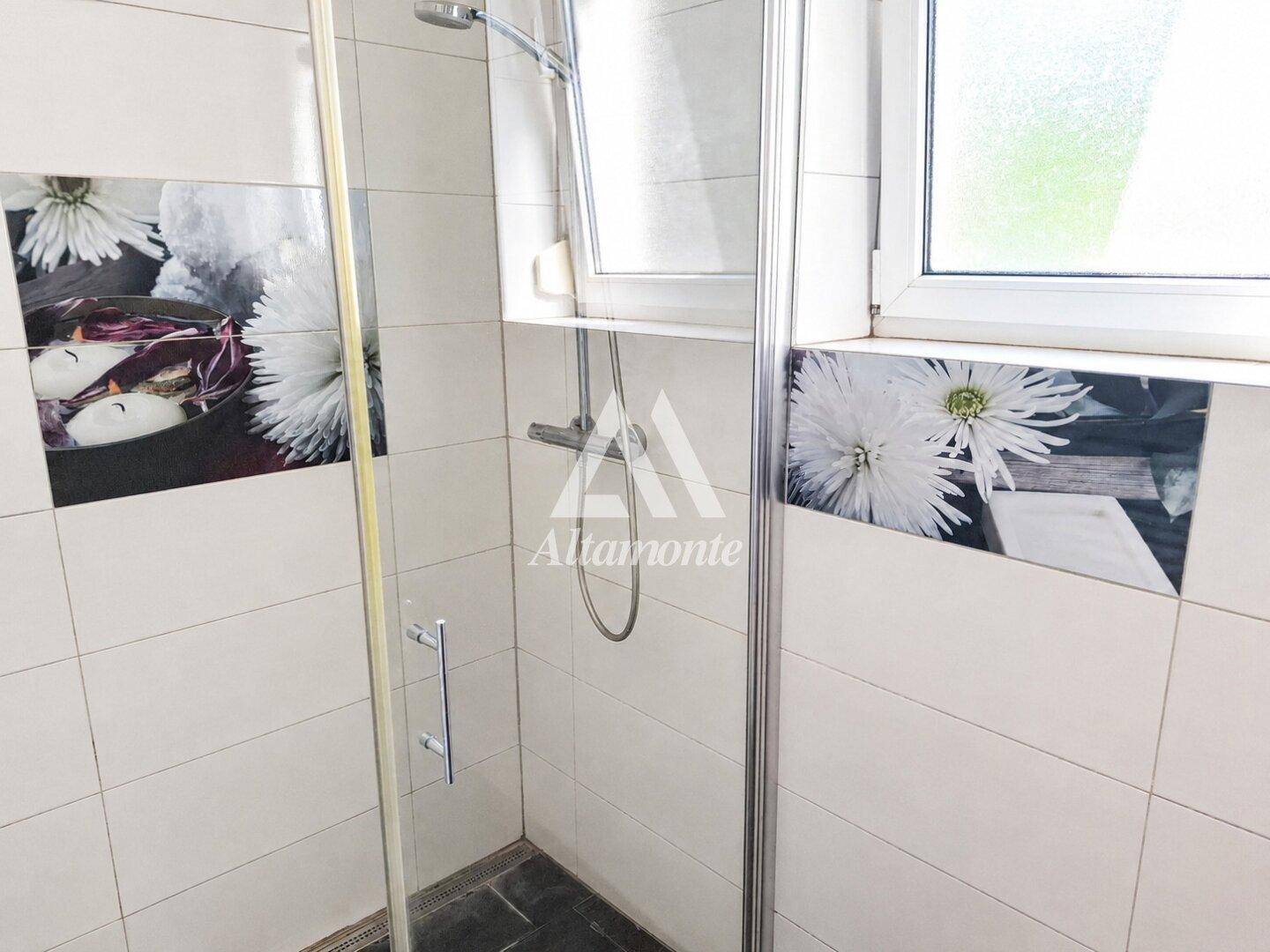 Duschbereich im Badezimmer mit weißen Fliesen, dekorativen Blumenmotiven und einem Fenster.