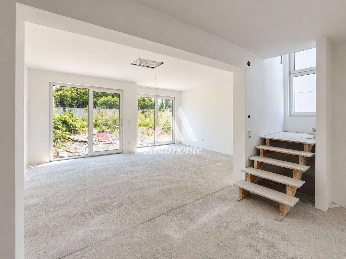 Geräumiges Wohnzimmer im Rohbauzustand mit Betonboden und großen Fenstern zum Garten.