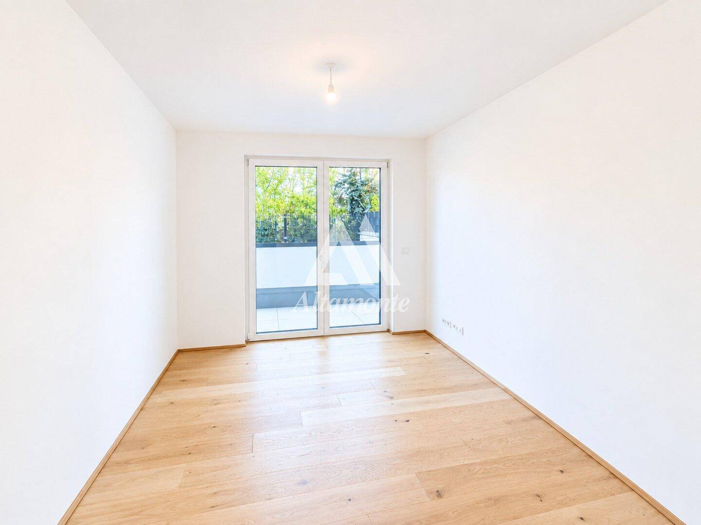 Leeres Zimmer mit hellem Holzboden und großen Fenstern, die Zugang zu einem Außenbereich bieten.