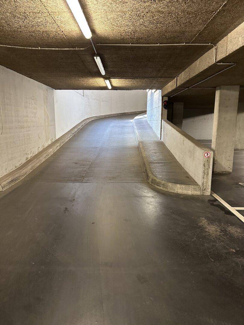 Zufahrtsrampe zu einer Tiefgarage mit Betonwänden und Beleuchtung, die in die Tiefe führt.