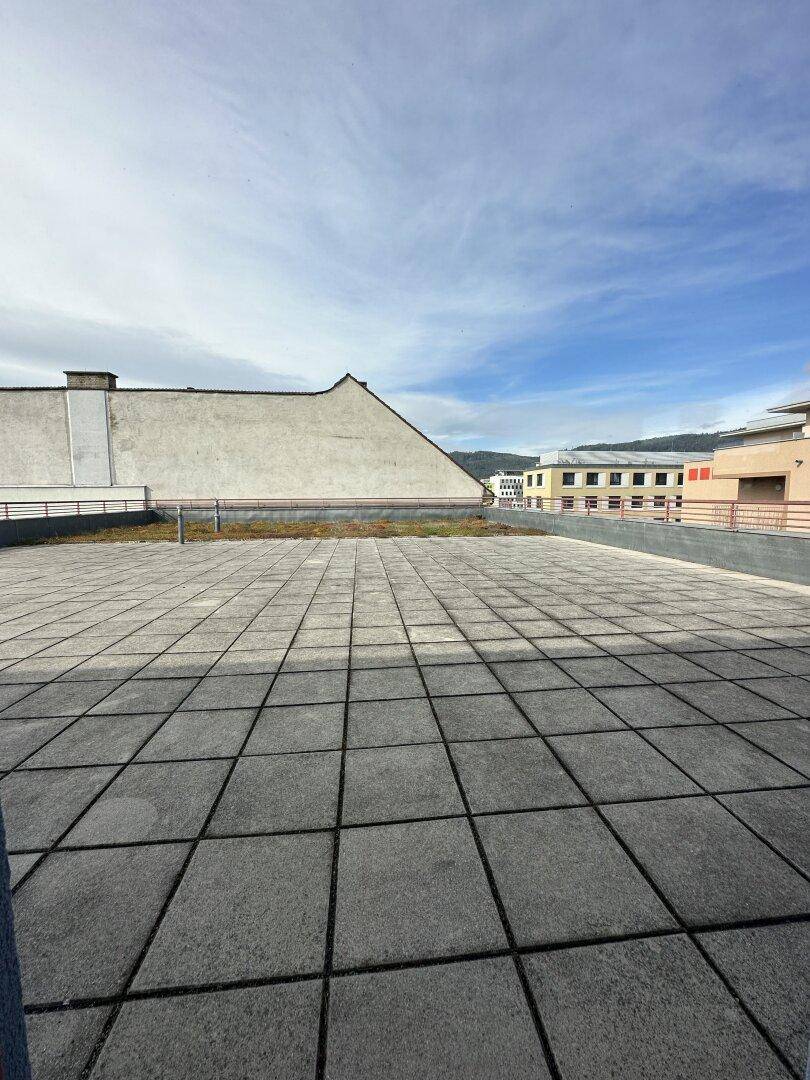 Weitläufige Dachterrasse mit grauen Fliesen und Blick auf die umliegenden Gebäude unter blauem Himmel.