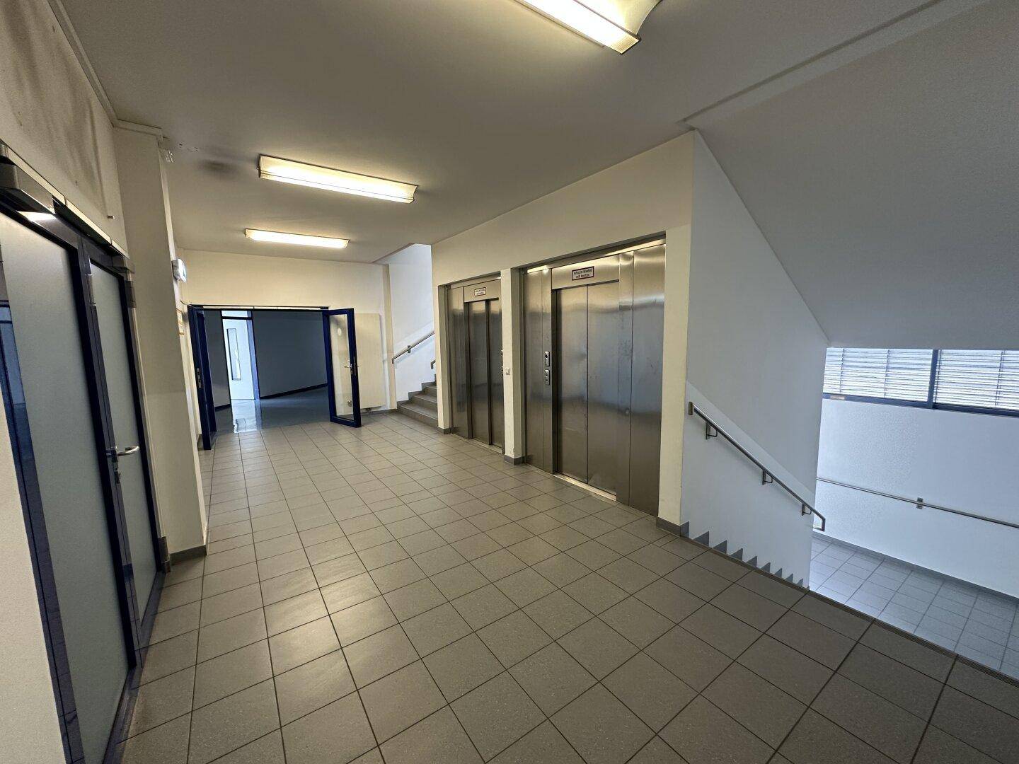 Geräumiger Flur mit zwei Aufzügen und einem Treppenhaus, ausgestattet mit grauen Fliesenböden.