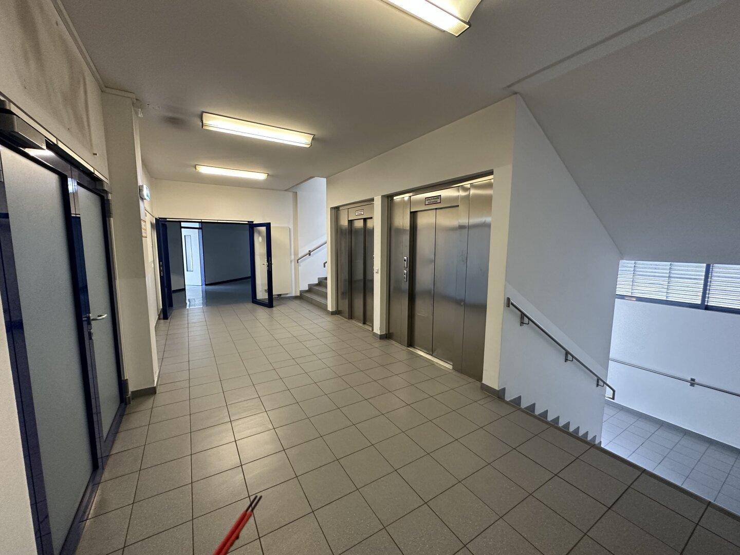 Geräumiger Flur mit zwei Aufzügen und einem Treppenhaus, ausgestattet mit grauen Fliesenböden.