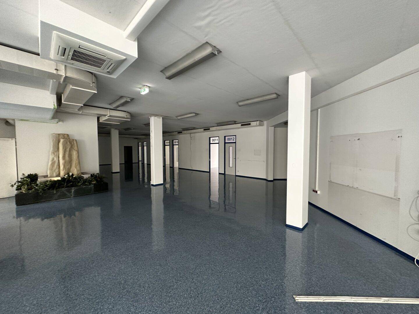 Großer, offener Büroraum mit glänzendem blauen Boden, Säulen und sichtbaren Lüftungsrohren an der Decke.