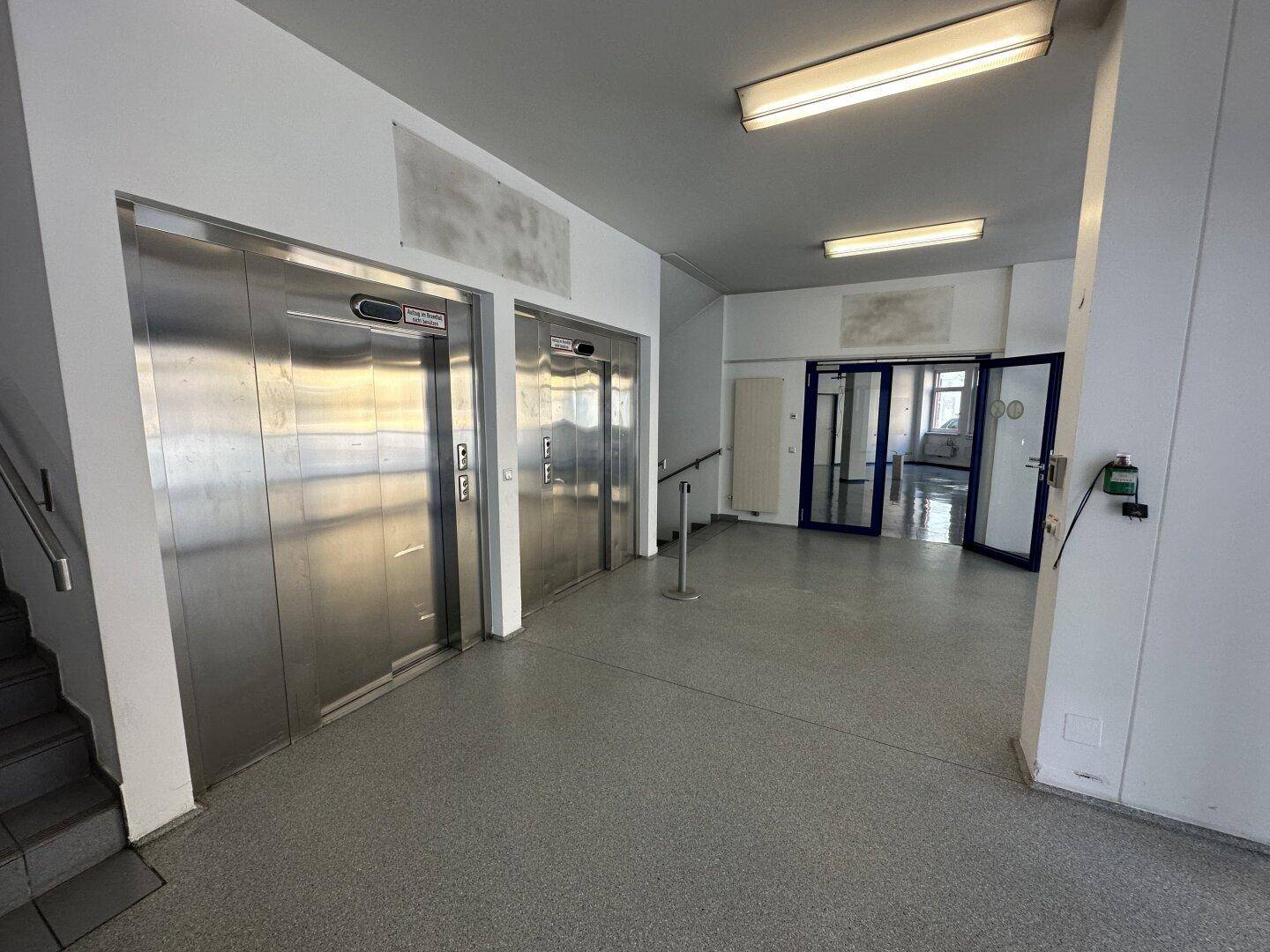 Heller Flur mit zwei Aufzügen und einem Treppenhaus, ausgestattet mit grauen Fliesenböden.