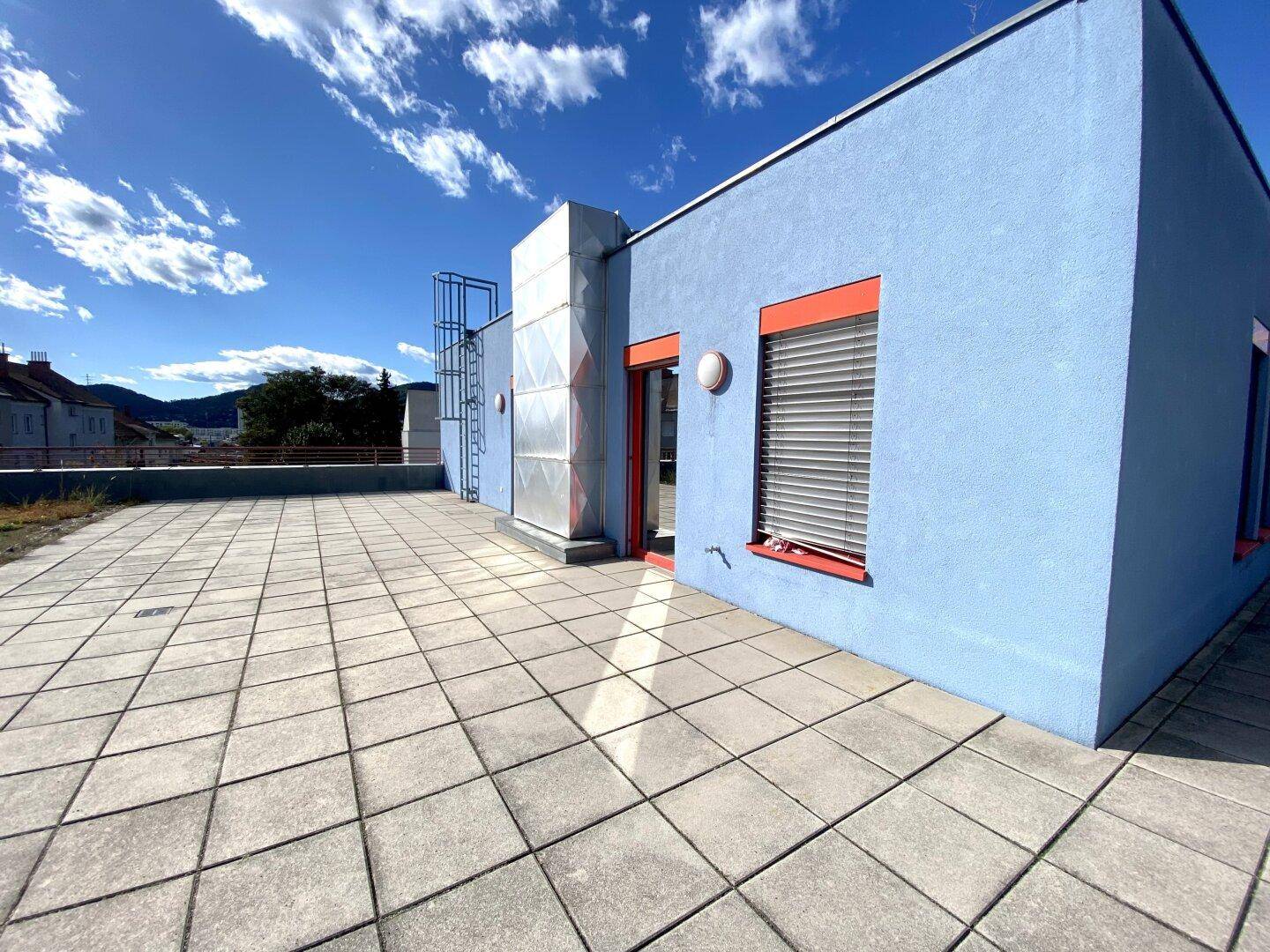 Weitläufige Dachterrasse mit grauen Fliesen und einem Zugang zum Gebäude unter blauem Himmel.