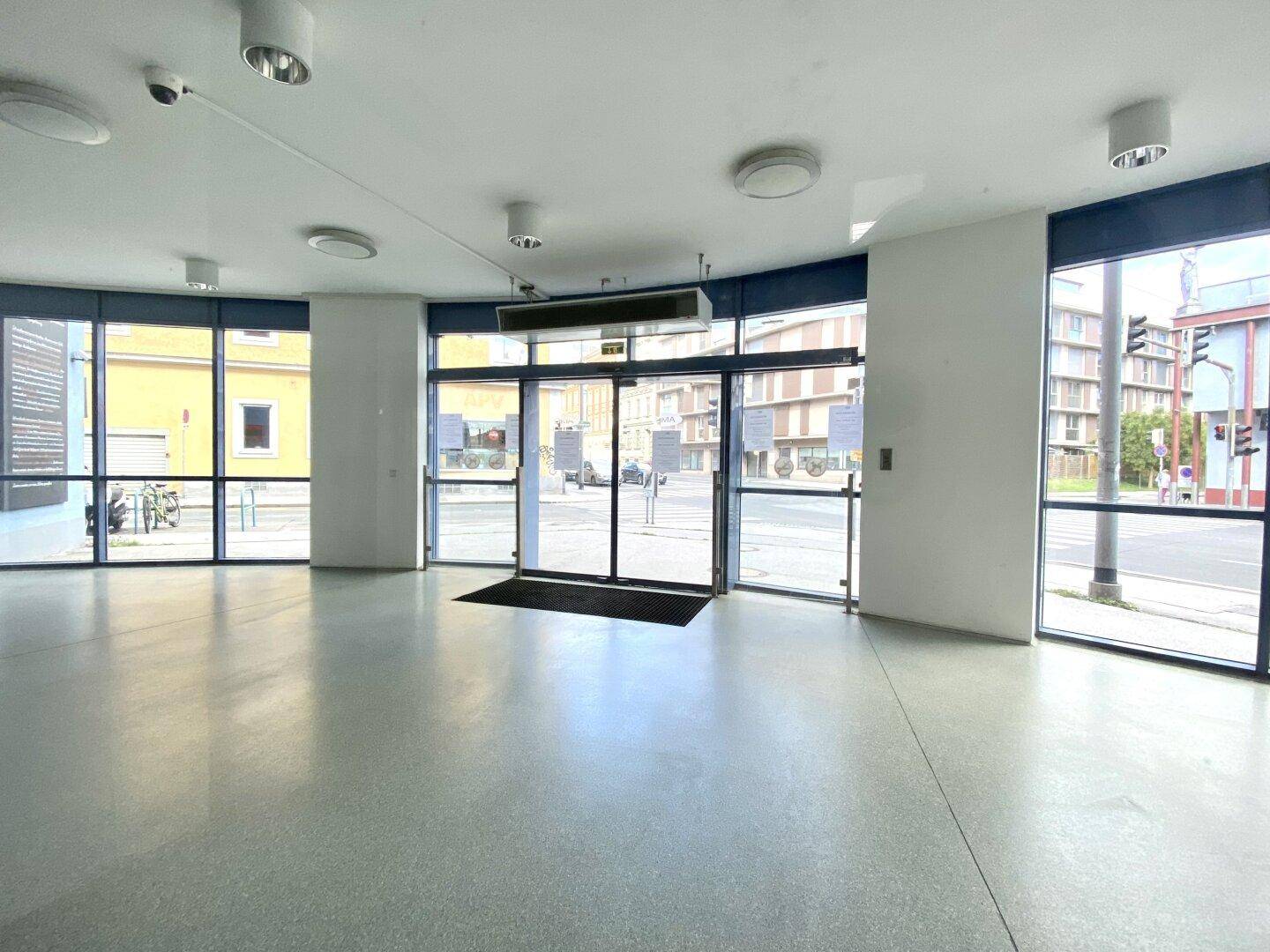Heller Eingangsbereich mit großen Glasfronten und automatischen Schiebetüren, die zur Straße führen.