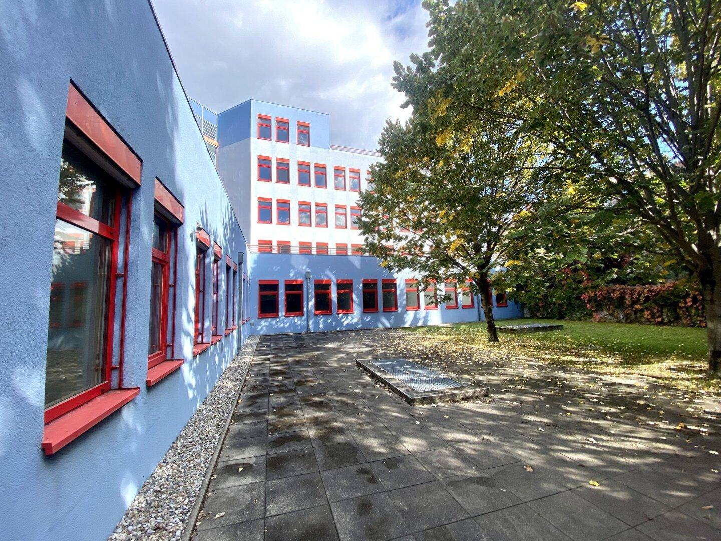 Grüner Innenhof mit Bäumen und gepflastertem Bereich, umgeben von einem Gebäude mit blauer Fassade.