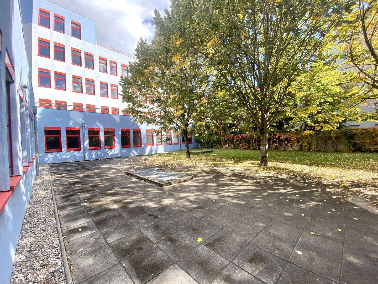 Grüner Innenhof mit Bäumen und gepflastertem Bereich, umgeben von einem Gebäude mit blauer Fassade.