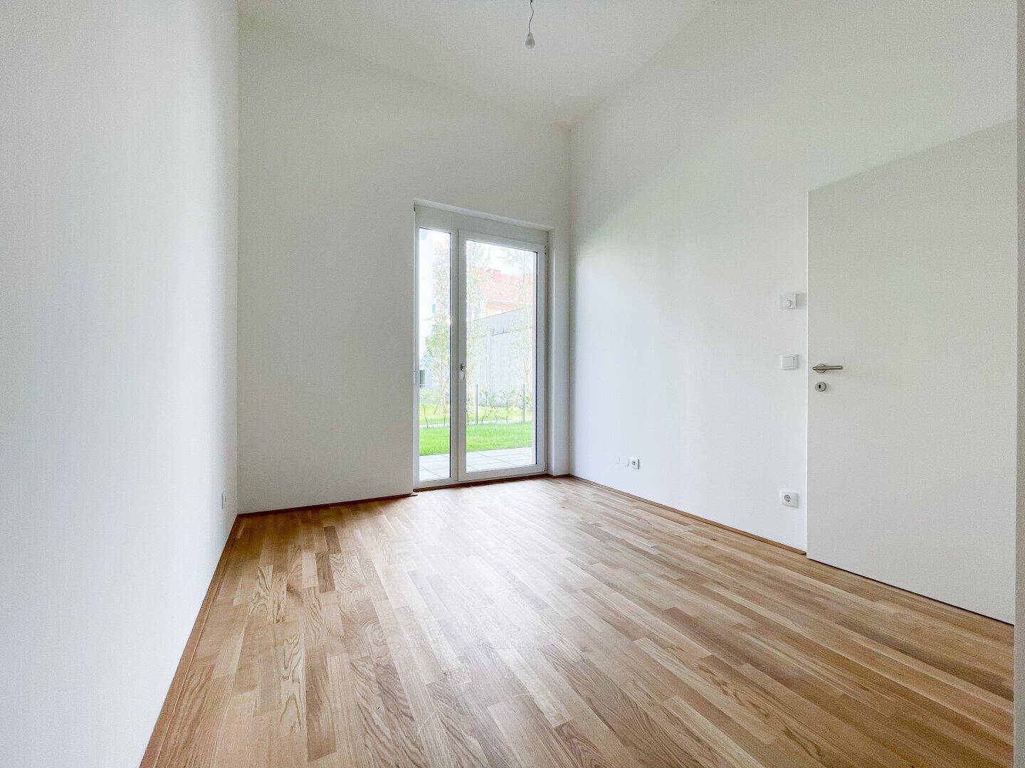 Leeres Zimmer mit hellem Holzparkettboden und Zugang zu einer Grünfläche.