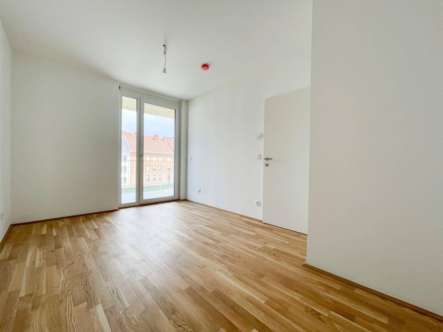 Heller Raum mit Holzboden und großem Fenster, das Zugang zu einem Balkon bietet.