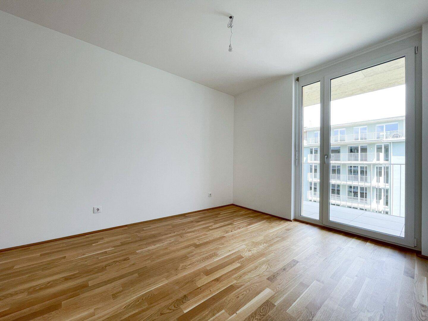 Geräumiger, heller Raum mit Holzboden und großen Fenstern mit Blick auf ein gegenüberliegendes Gebäude.