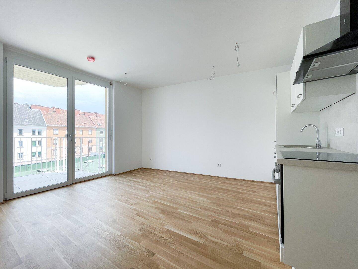 Heller Wohnbereich mit Holzboden und Zugang zu einem Balkon, angrenzend an eine Küchenzeile.