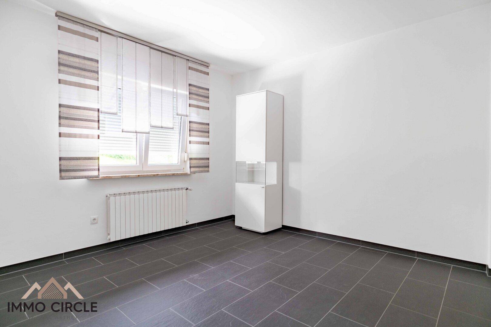 Leeres Zimmer mit dunklen Fliesen, großem Fenster und Heizkörper.