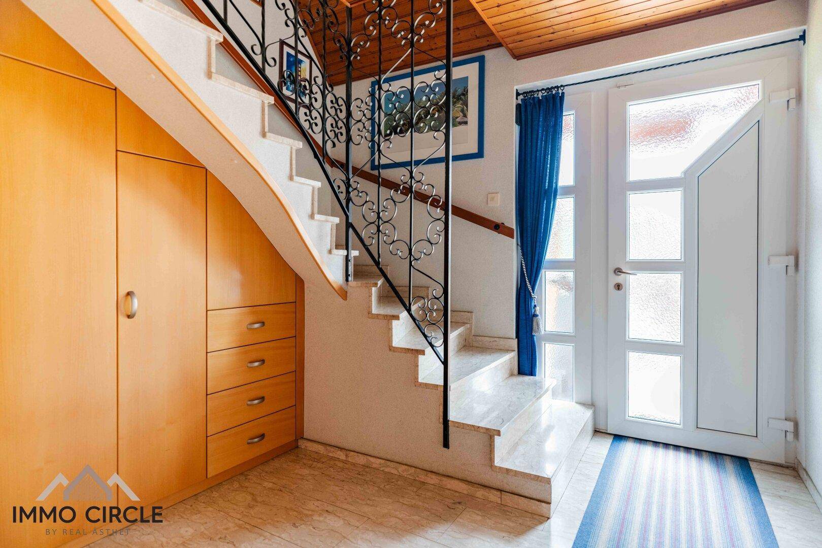 Heller Eingangsbereich mit Marmortreppe, schmiedeeisernem Geländer und Holzdecke.