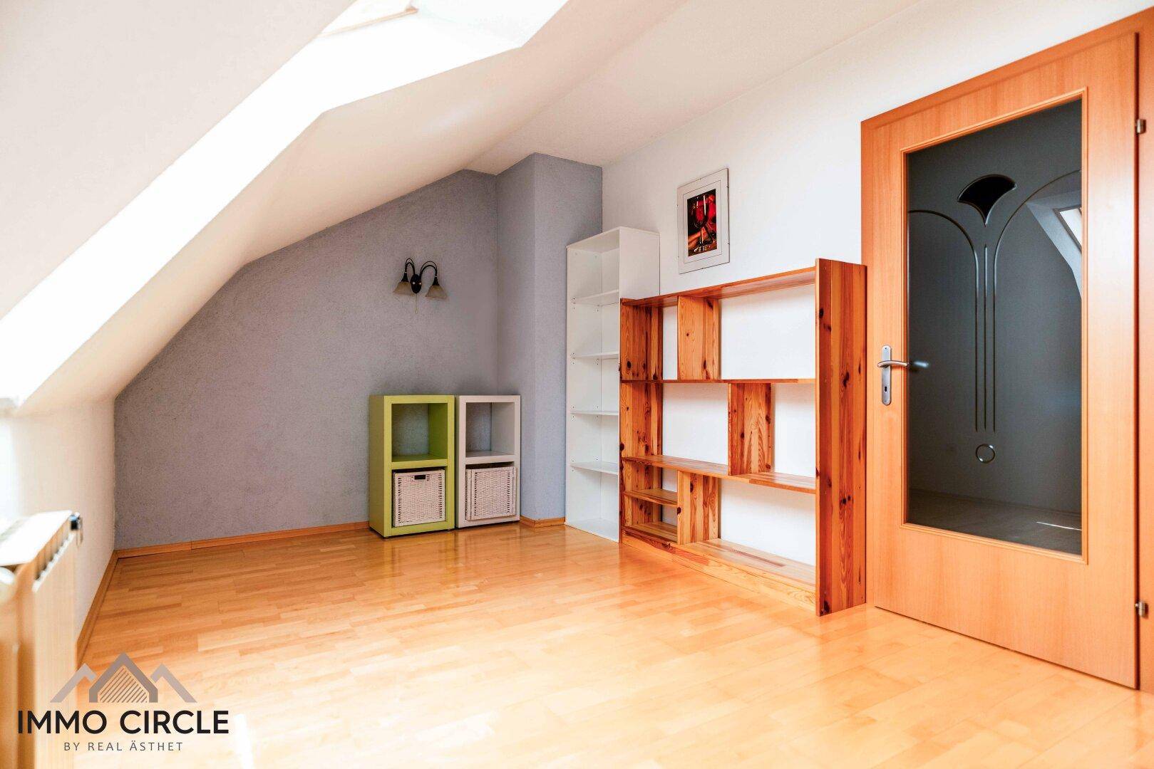 Helles Zimmer im Dachgeschoss mit Holzboden, Dachschräge und Regalen.