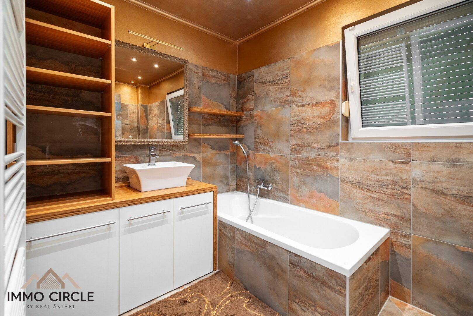 Helles Badezimmer mit Badewanne, Fenster und gemusterten Wand- und Bodenfliesen.