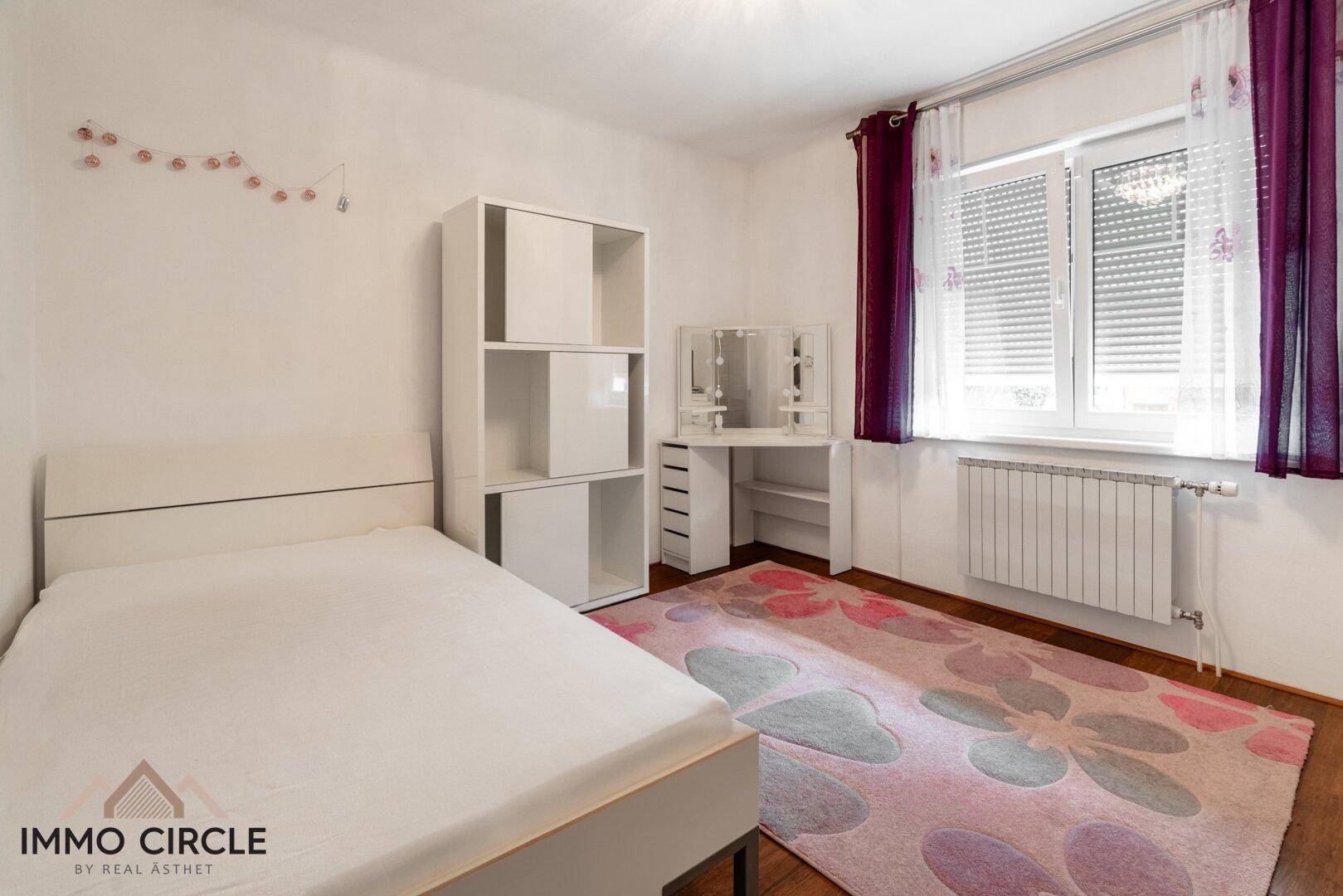 Helles Schlafzimmer mit weißem Bett, Schminktisch und farbenfrohem Teppich vor dem Fenster.
