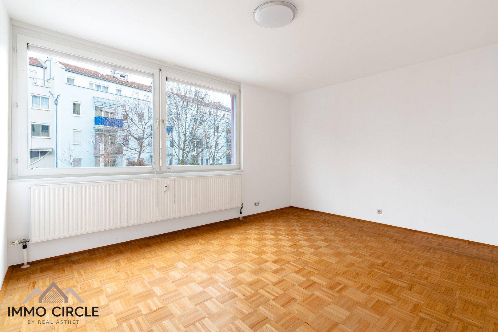 Helles Zimmer mit Parkettboden und einem großen Fenster mit Blick nach draußen.