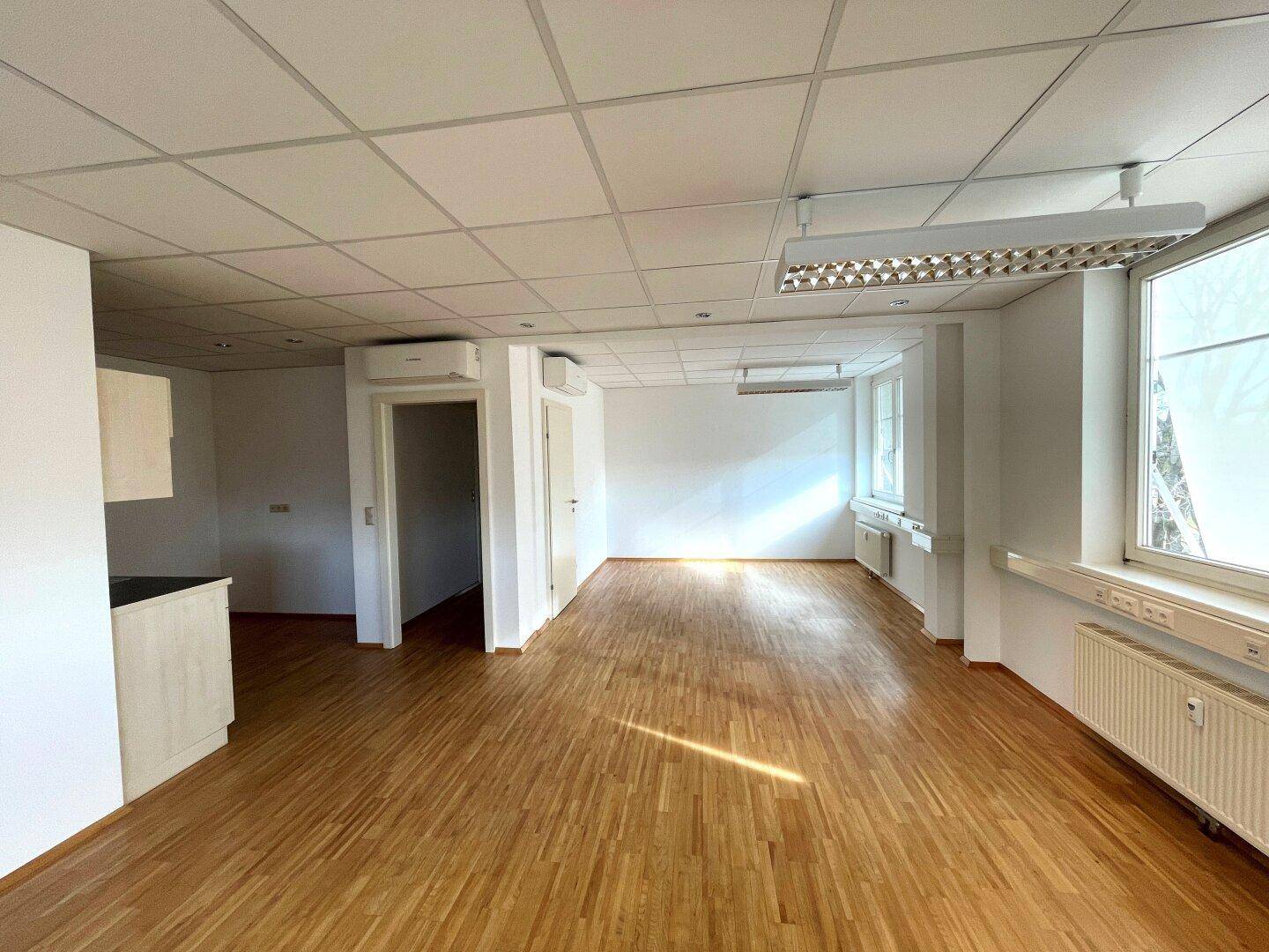 Helles Büro mit Holzparkettboden, abgehängter Decke und einem kleinen Küchenbereich für Mitarbeiter.