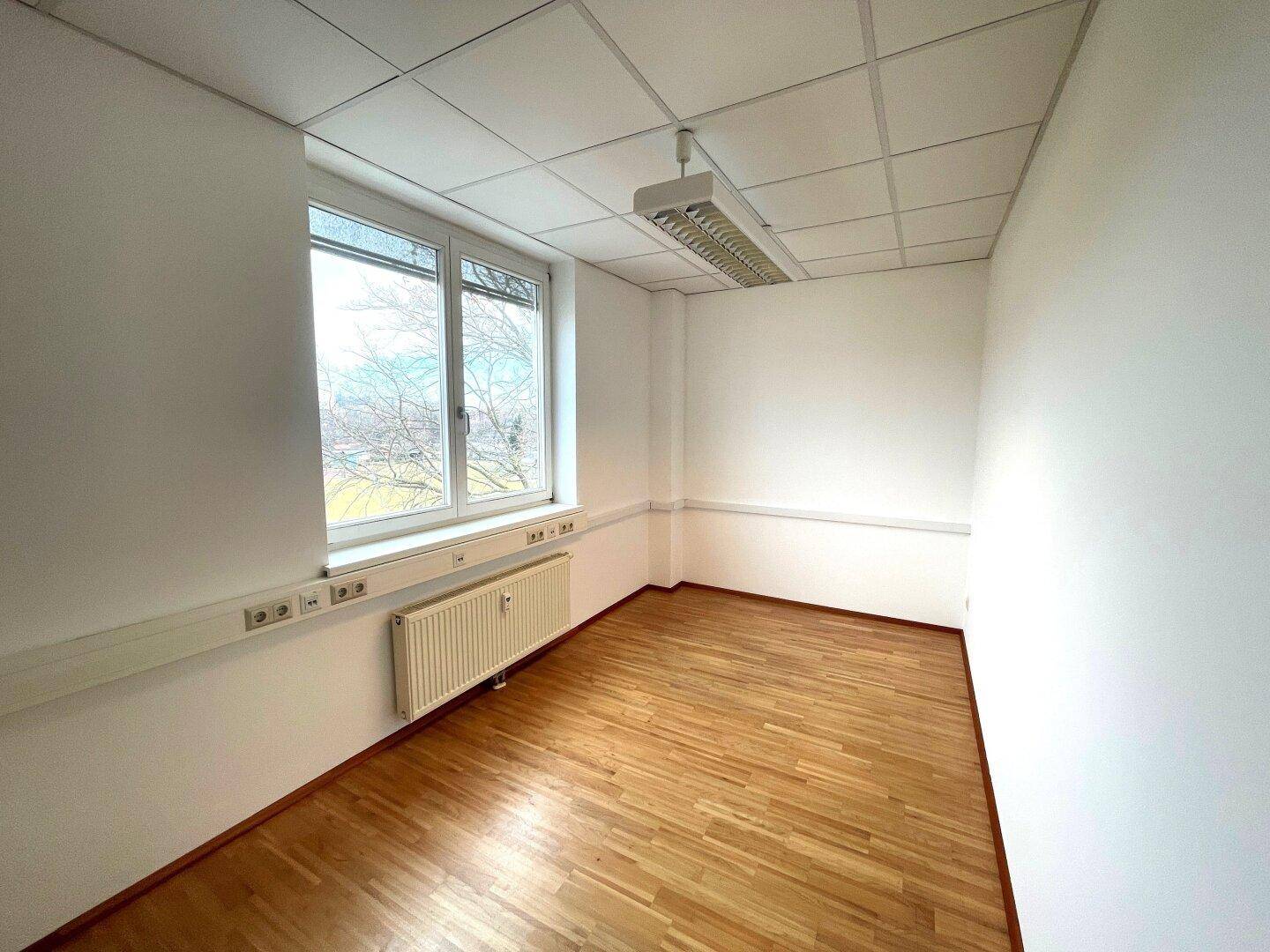 Kleines, helles Büro mit Holzparkettboden, einem Fenster und weiß gestrichenen Wänden, ideal für Einzelarbeit.