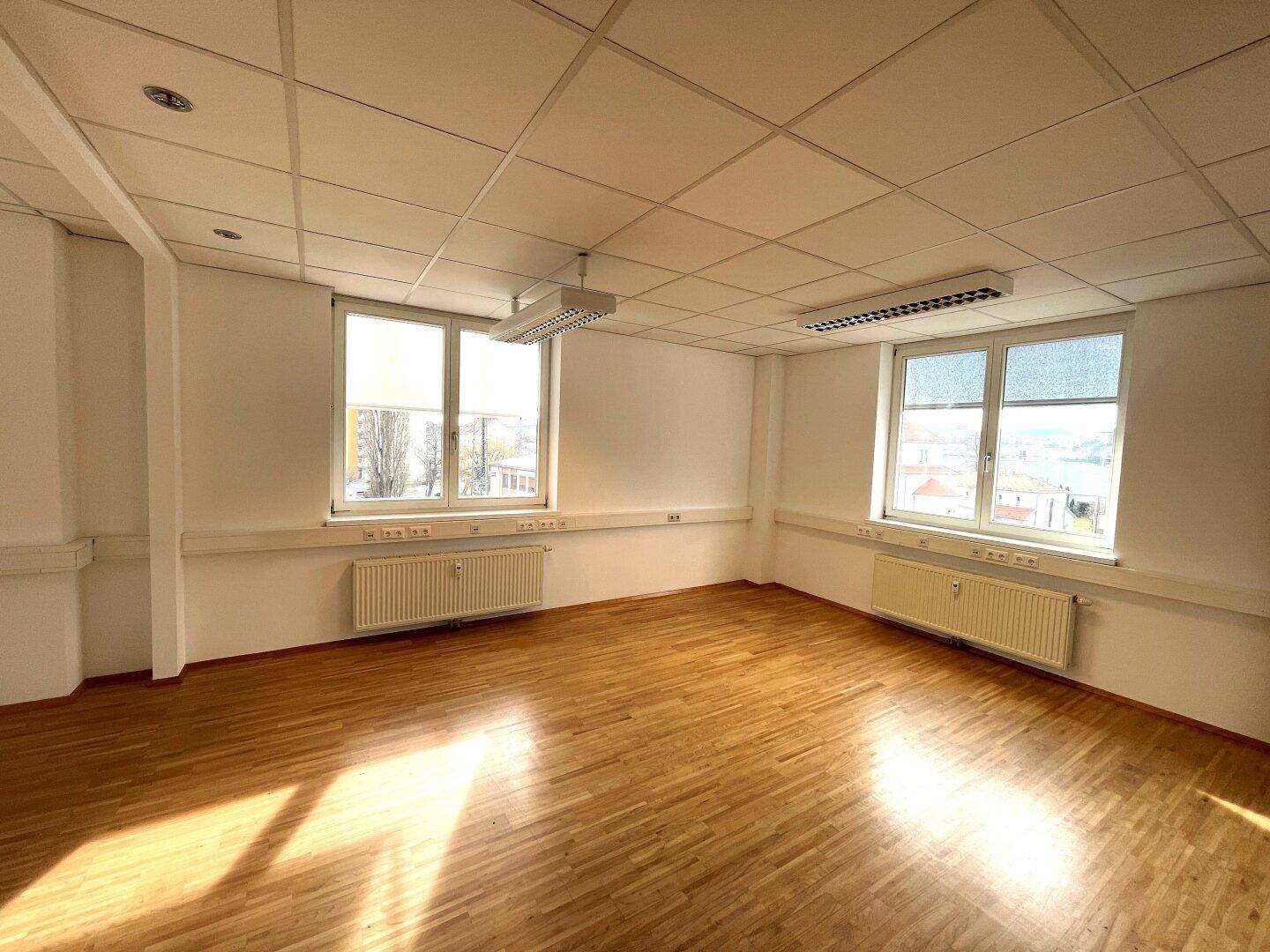 Leeres Büro mit Holzparkettboden, großen Fenstern und abgehängter Decke, gut beleuchtet durch Tageslicht.