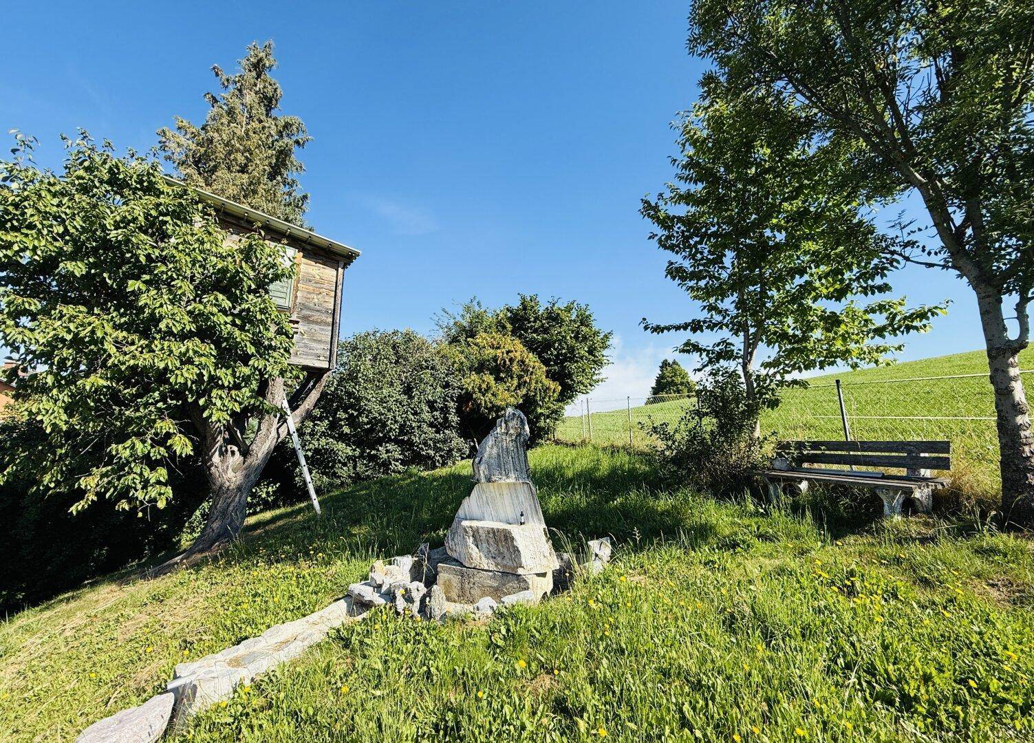 Idyllischer Gartenbereich mit Baumhaus, Sitzbank und einem kleinen Wasserlauf in grüner Natur.