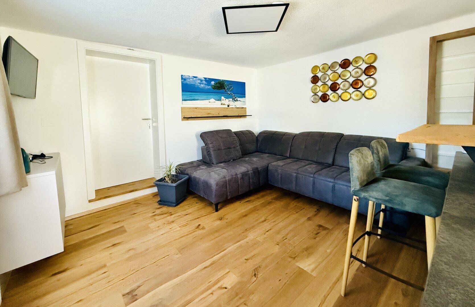 Gemütlicher Wohnbereich mit Ecksofa, Barhockern und Holzboden in hellem Raum.
