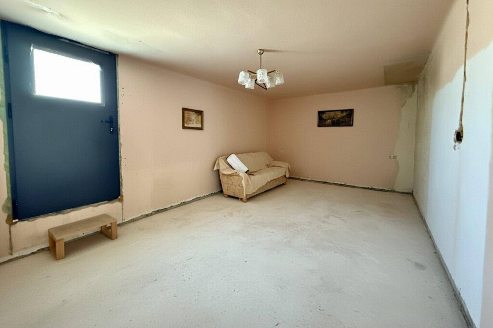 Unfertiger Wohnraum mit einfacher Couch, Kronleuchter und Renovierungsbedarf.