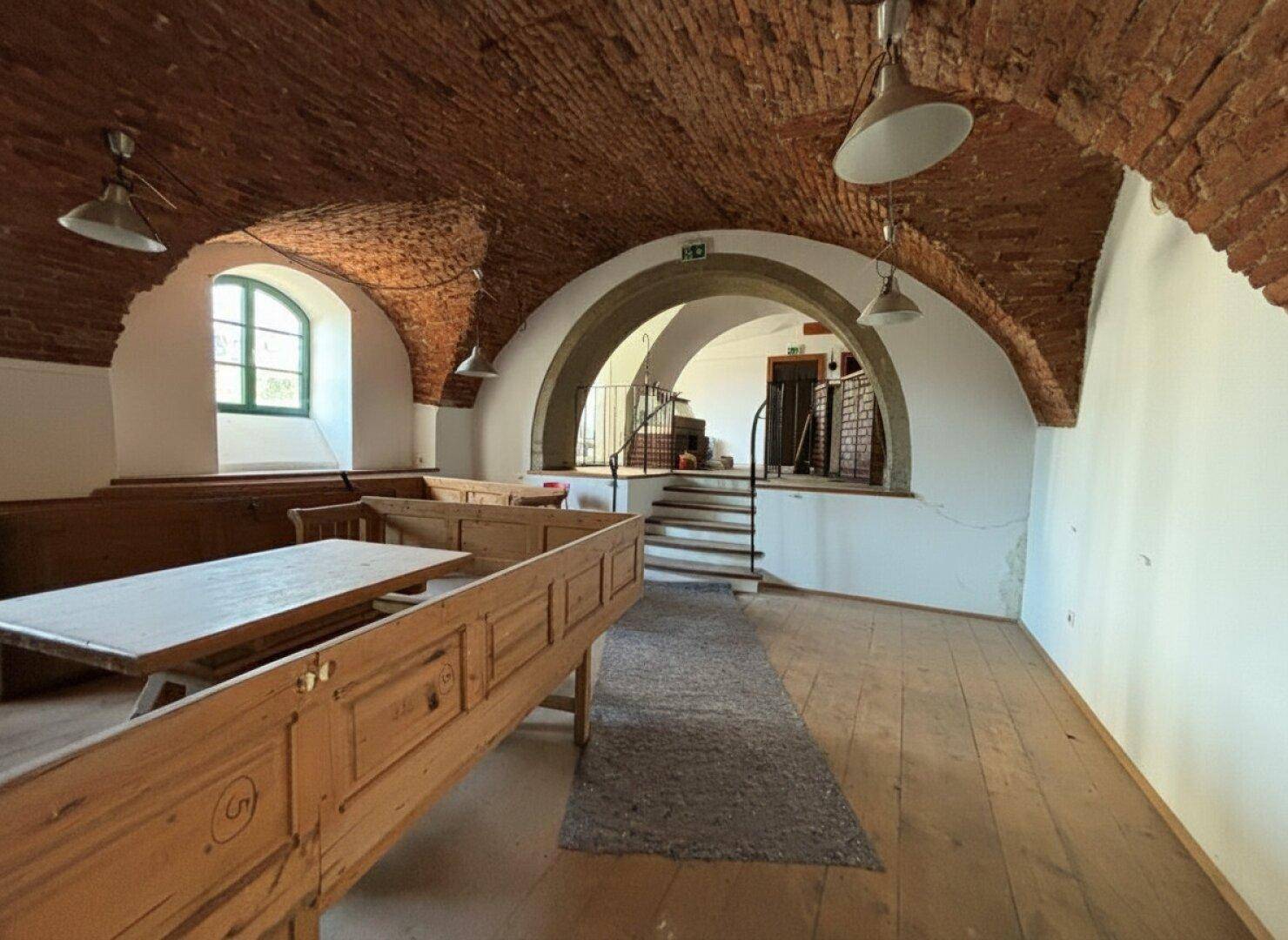 Geräumiger Gastraum mit Holzbänken, Ziegelgewölbedecken und einem Übergang zu einem erhöhten Bereich, der renoviert werden muss.