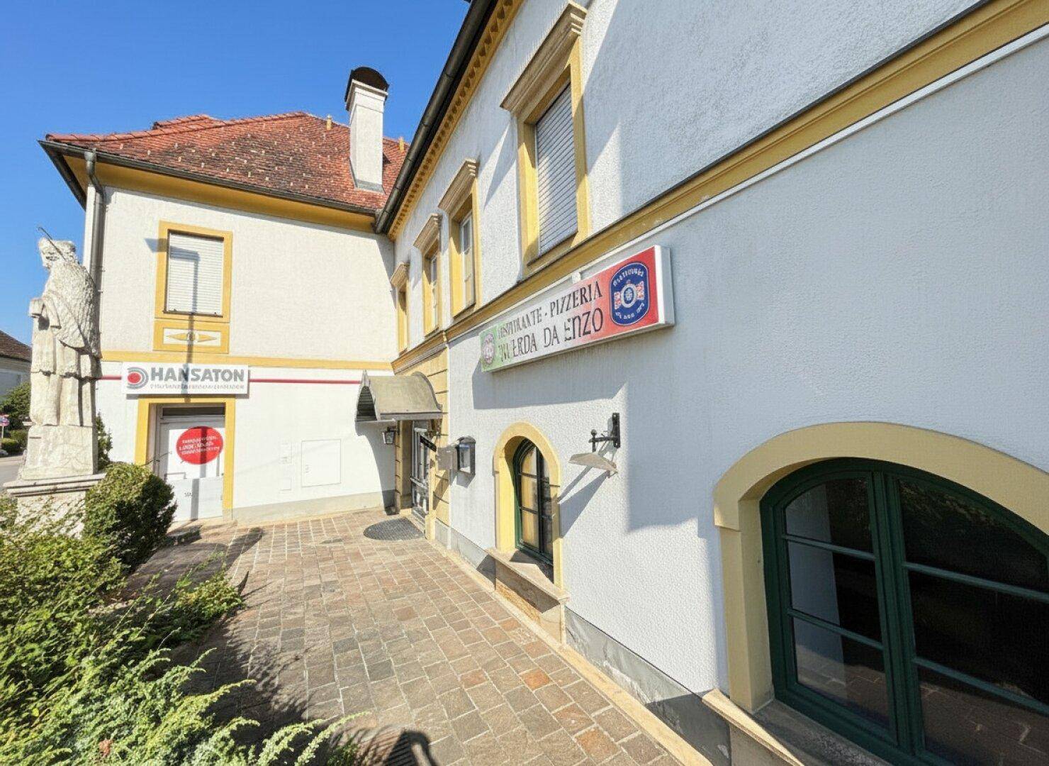 Seitenansicht des Gebäudes mit Restaurantbeschilderung, gepflastertem Weg und einer Statue im Vordergrund bei Sonnenschein.