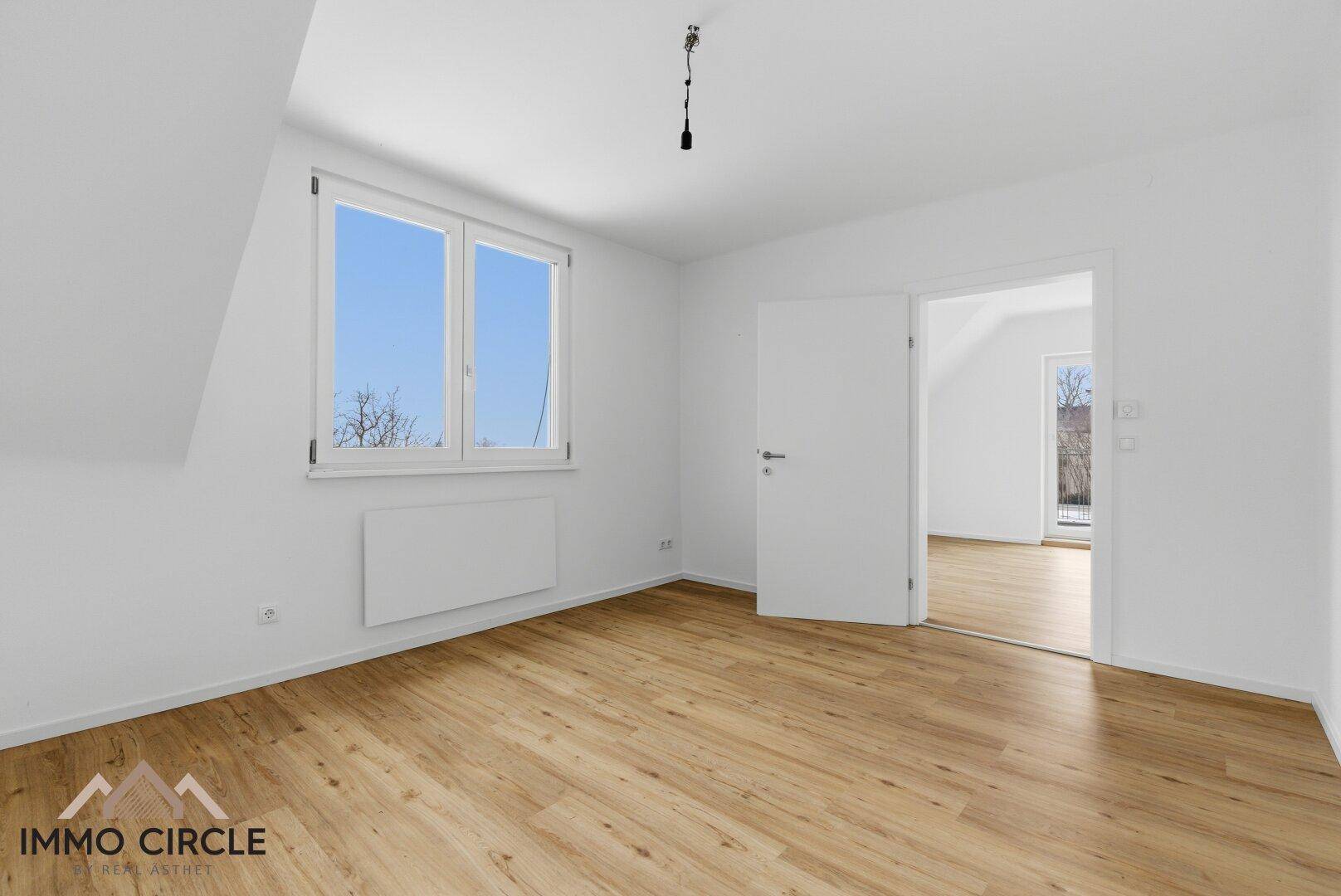 Geräumiges Zimmer mit Holzboden und weiß gestrichenen Wänden, bietet Zugang zu einem weiteren Raum.