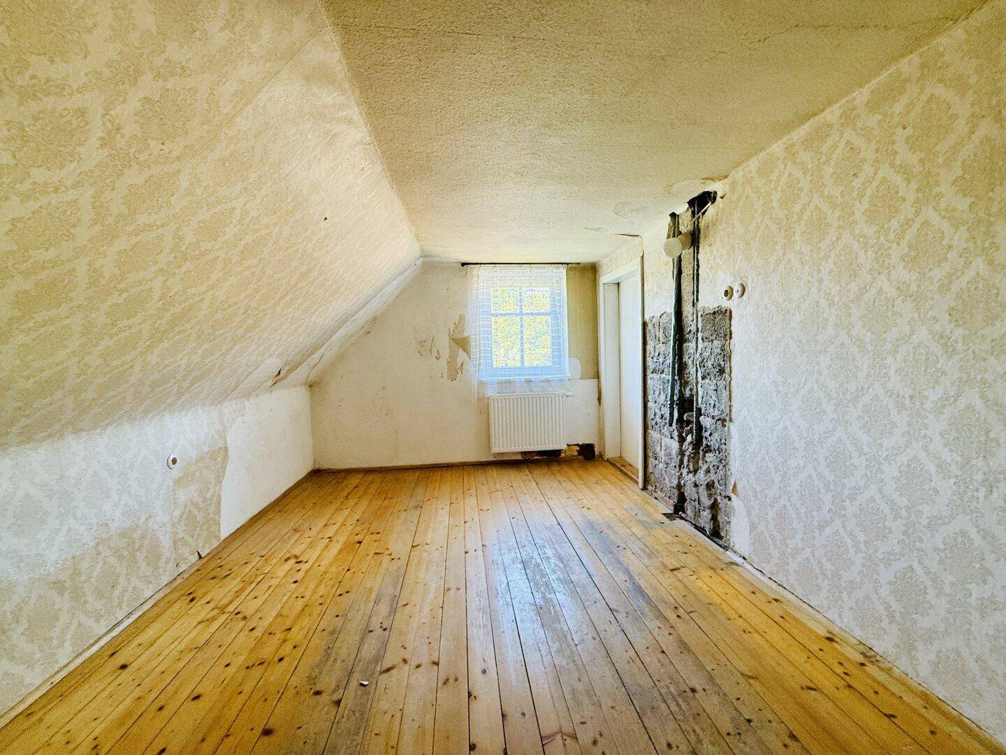 Dachgeschossraum mit Holzdielenboden, Fenster und abblätternder Tapete, renovierungsbedürftig.