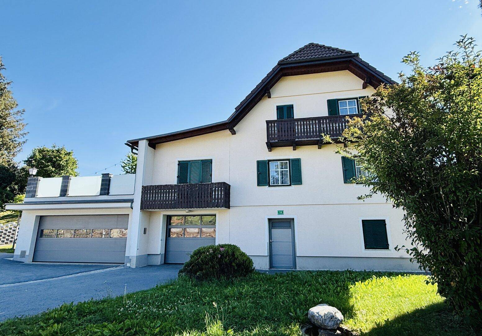 Wohnhaus mit weißer Fassade, zwei Garagen und traditionellen Holzbalkonen in ländlicher Umgebung.