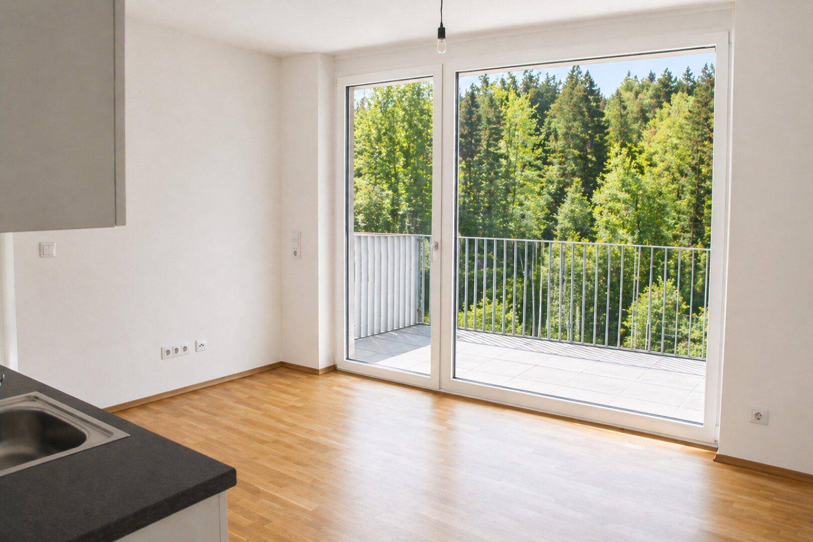 Heller Wohnbereich mit Holzboden, großer Fensterfront und Zugang zum Balkon mit Waldblick.