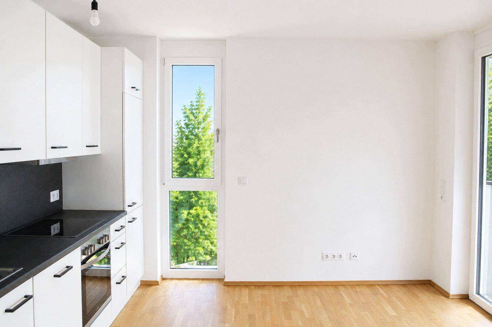 Moderne Einbauküche mit weißen Schränken, dunkler Arbeitsplatte und Fenster mit Waldblick.