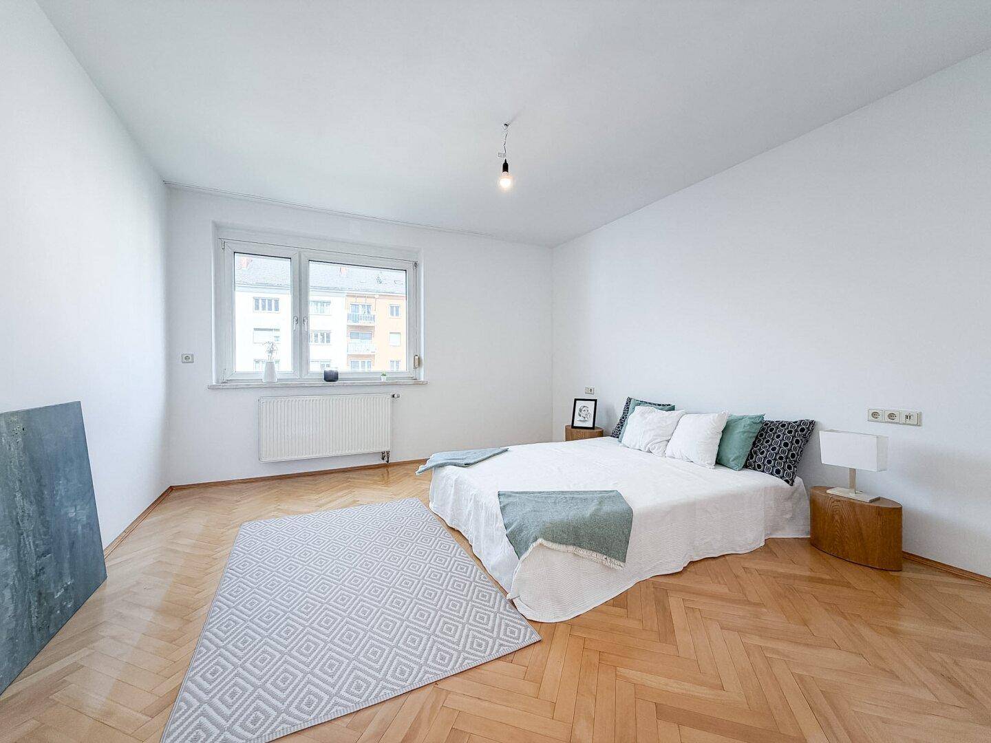 Schlafzimmer mit hellem Fischgrätparkett, großem Fenster und modernem Bett.