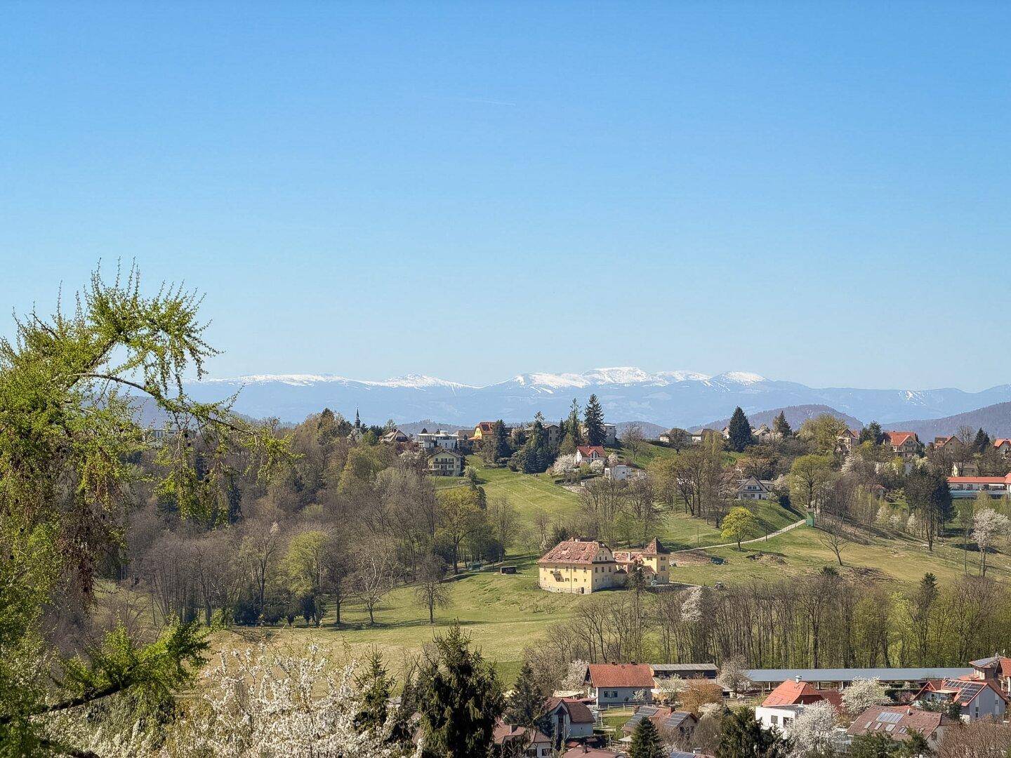 Panoramablick über eine hügelige Landschaft mit Häusern und schneebedeckten Bergen im Hintergrund.