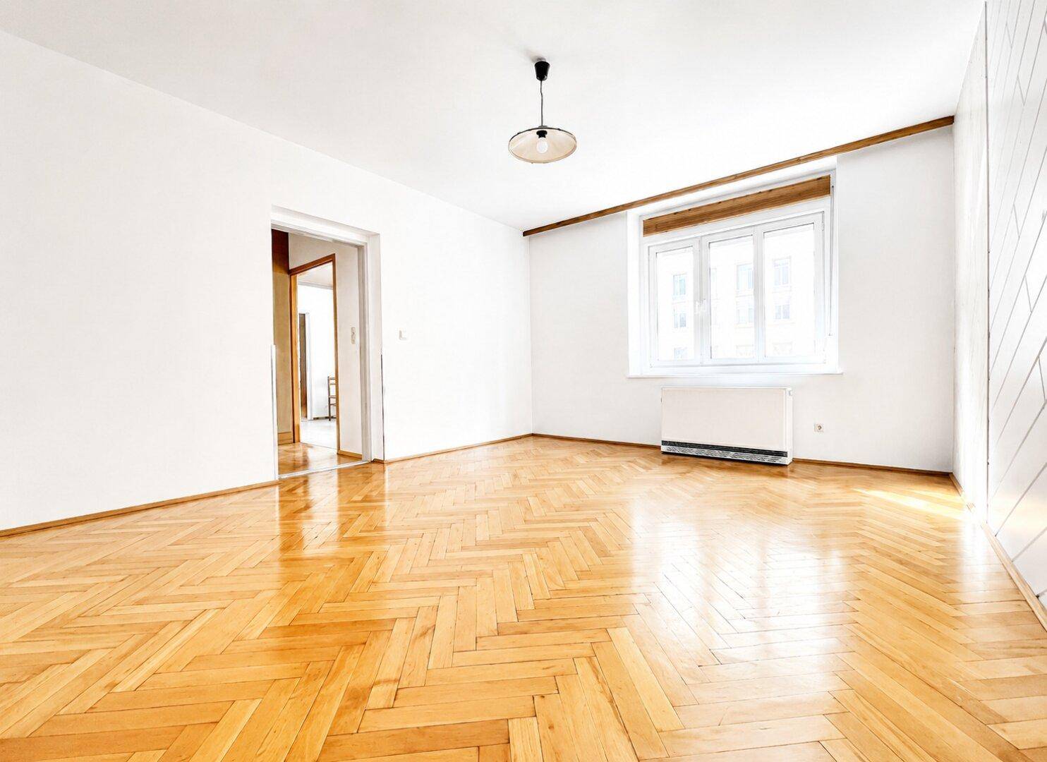 Geräumiges Wohnzimmer mit Fischgrätparkettboden und einem großen Fenster, das viel Licht hereinlässt.