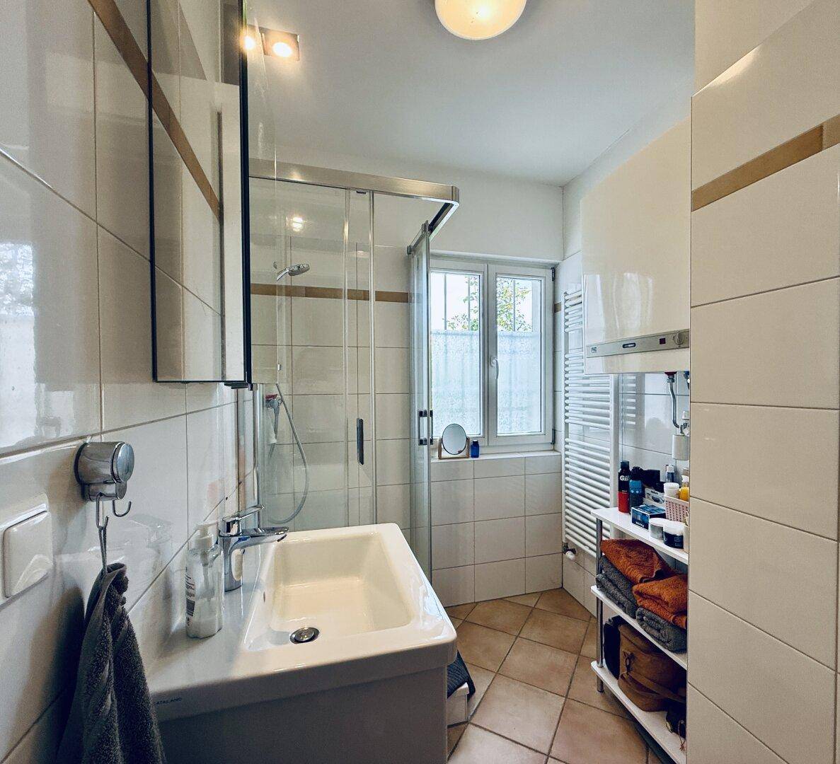 Kleines Badezimmer mit Duschkabine, Waschbecken, Fenster und weiß gefliesten Wänden.