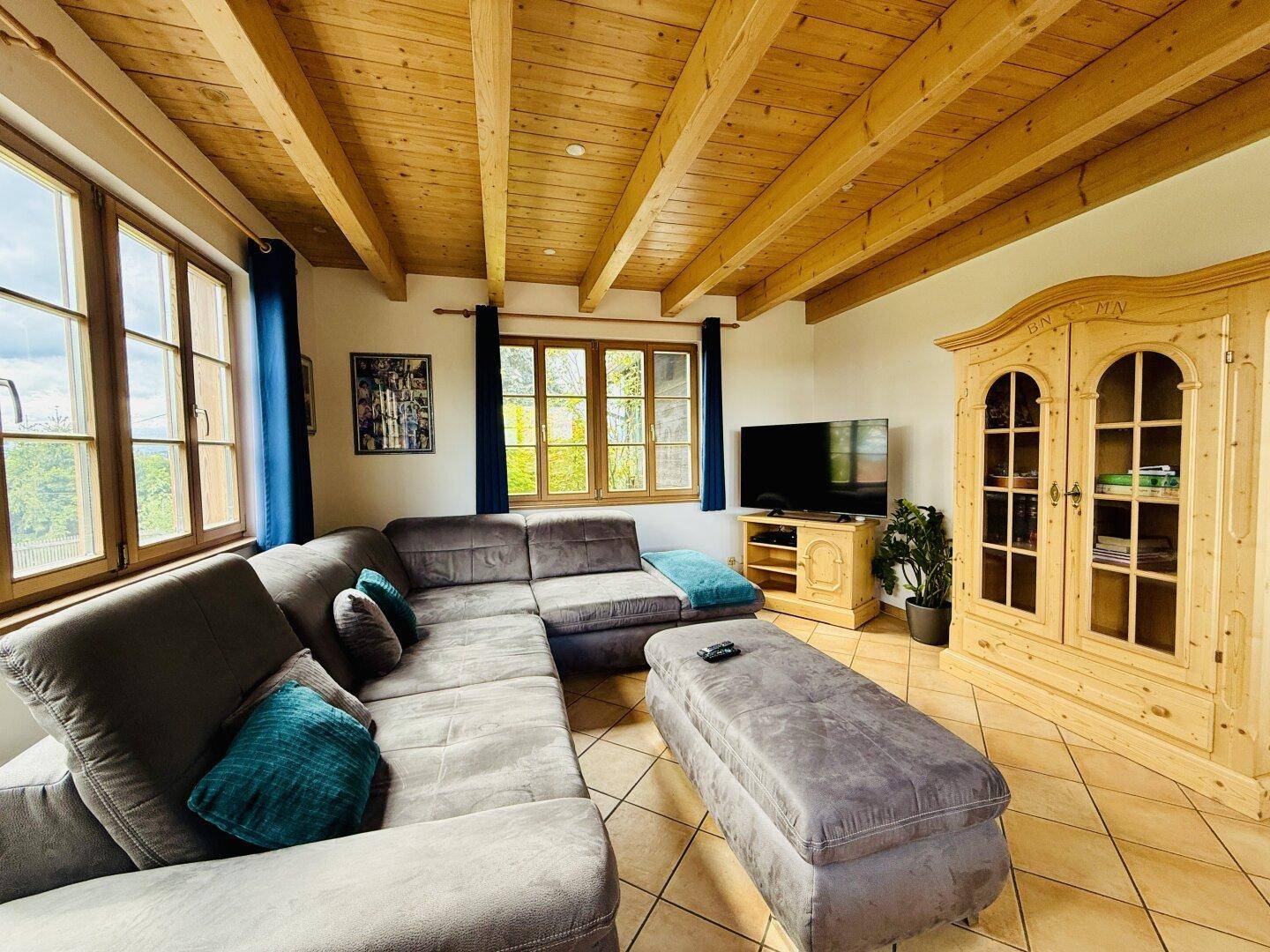Helles Wohnzimmer mit sichtbaren Holzbalken, großem Ecksofa und Fliesenboden.