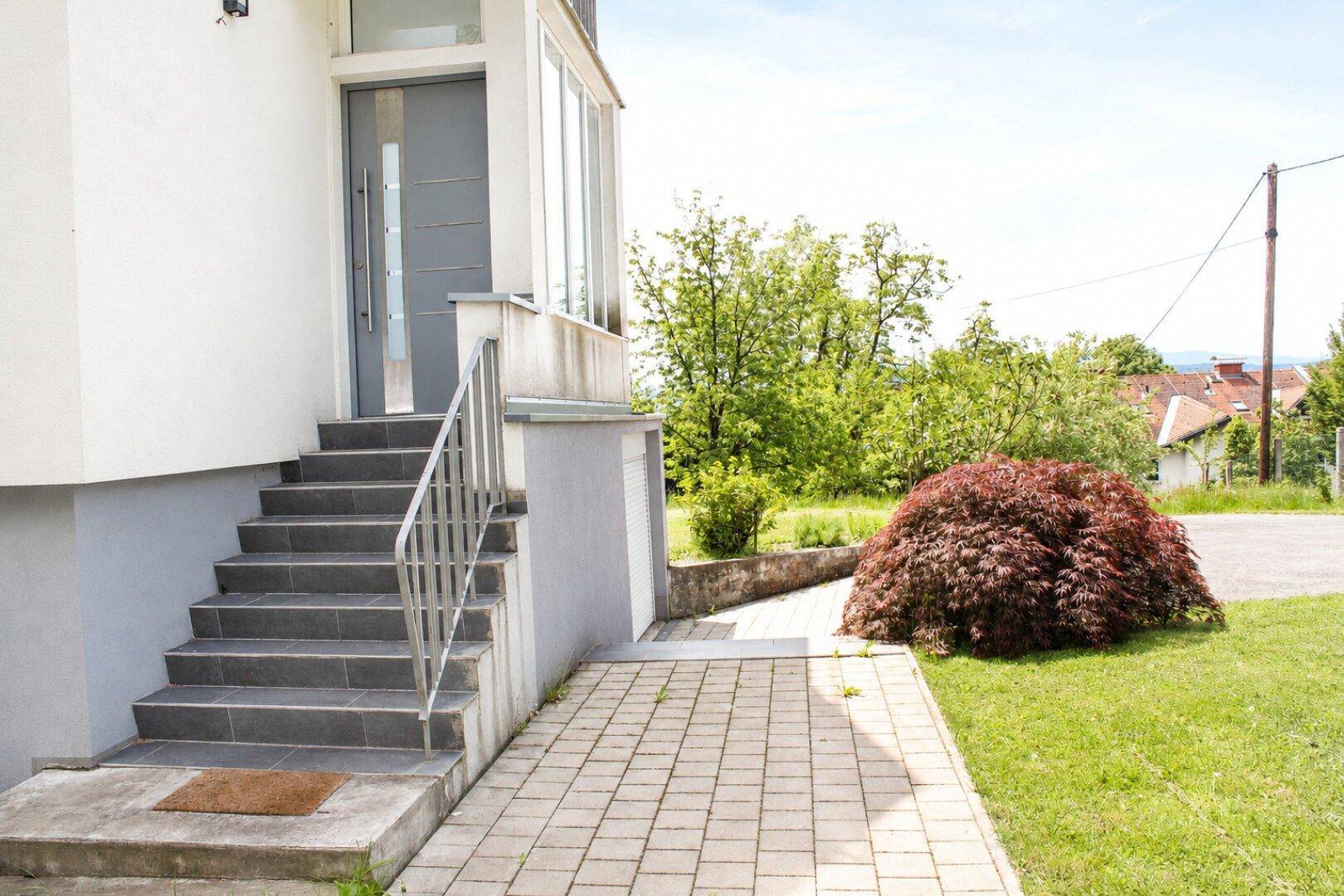 Eingangsbereich des Hauses mit moderner grauer Tür, Steintreppe und gepflastertem Zugangsweg.