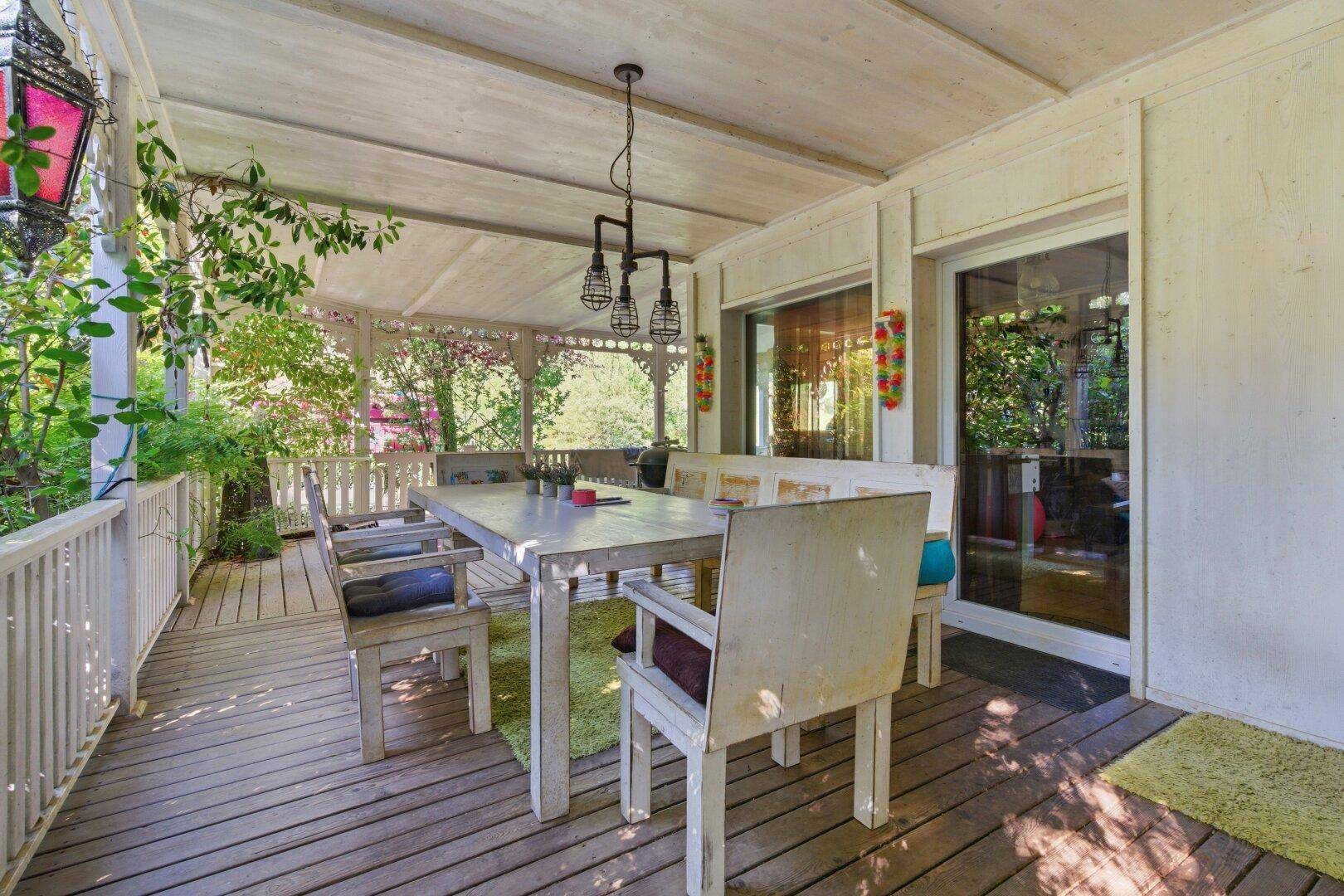 Überdachte Holzterrasse mit Esstisch und Stühlen, umgeben von üppigem Grün.