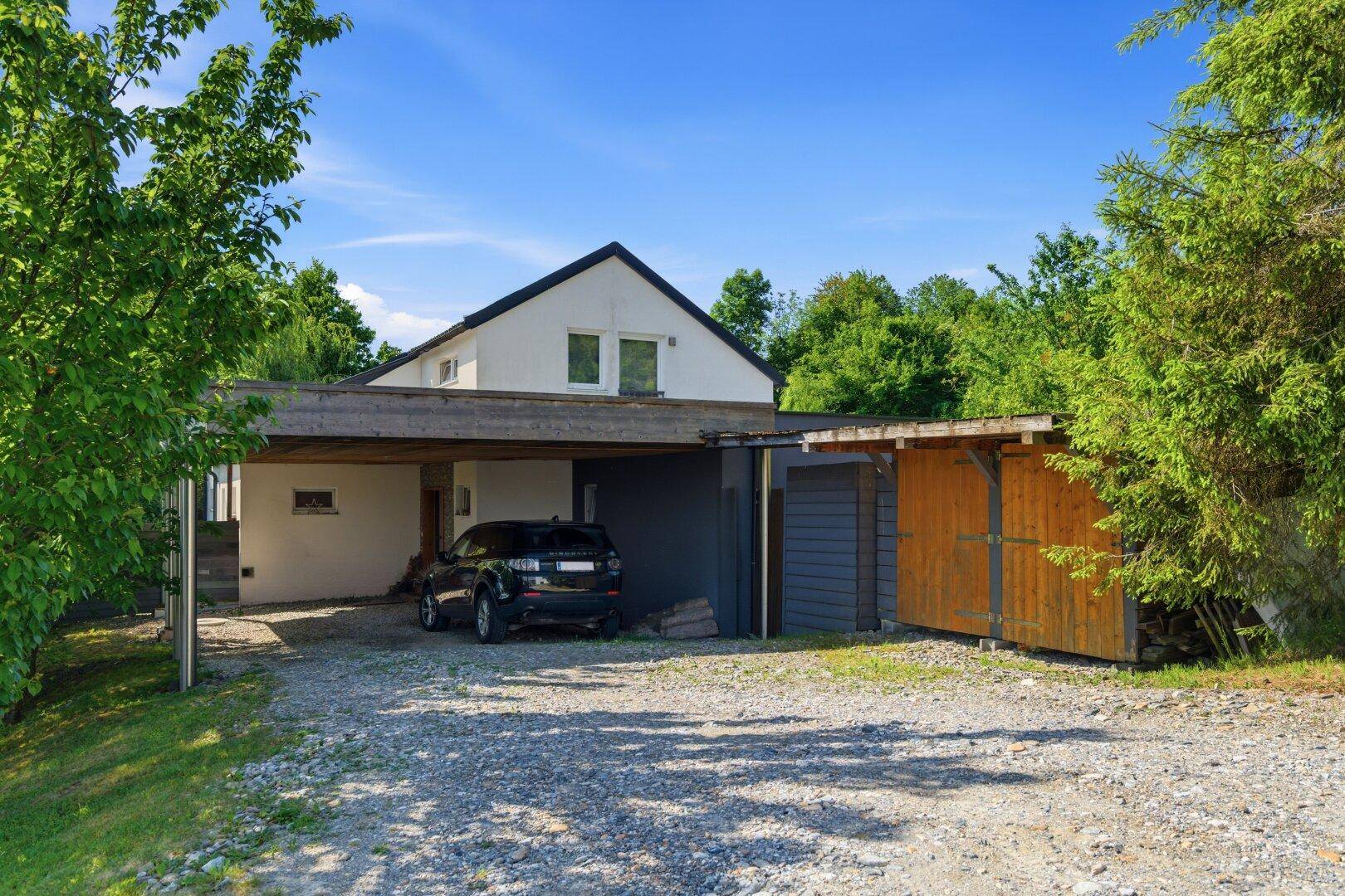 Vorderansicht des Hauses mit Carport, Schuppen und Kiesauffahrt in ländlicher Umgebung.