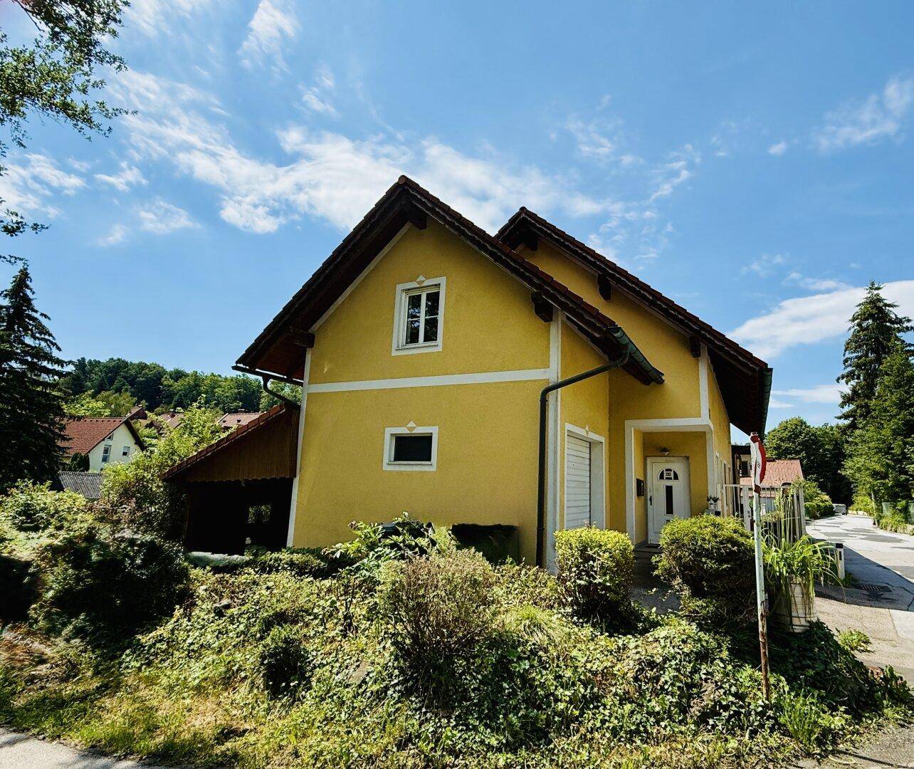 Gelbes Haus mit rotem Dach, umgeben von üppigem Grün und Bäumen unter blauem Himmel.