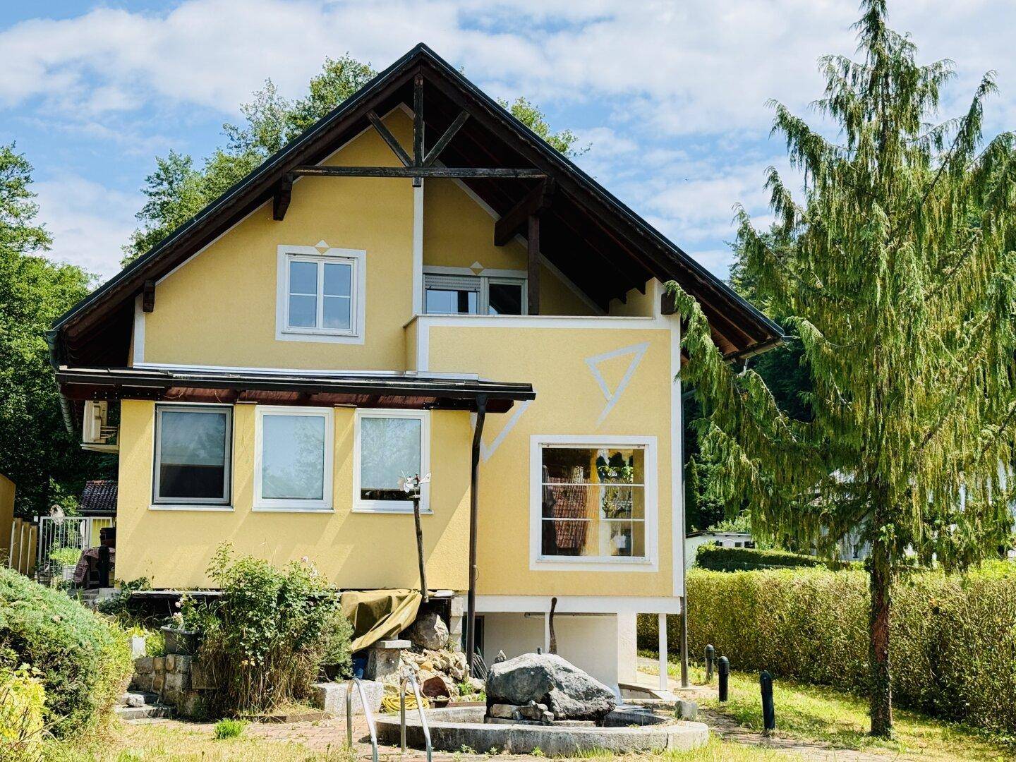 Gelbes Haus mit Satteldach und Balkon, umgeben von Bäumen und einem gepflegten Gartenbereich.