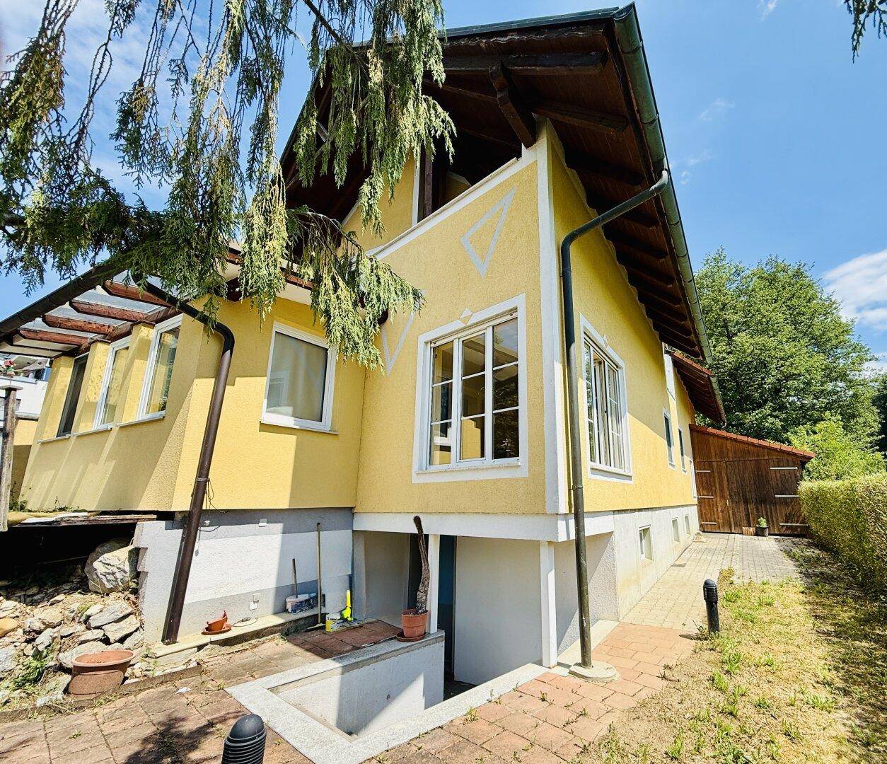 Seitenansicht des gelben Hauses mit Kellerzugang und einem Bereich mit Bäumen und Sträuchern.